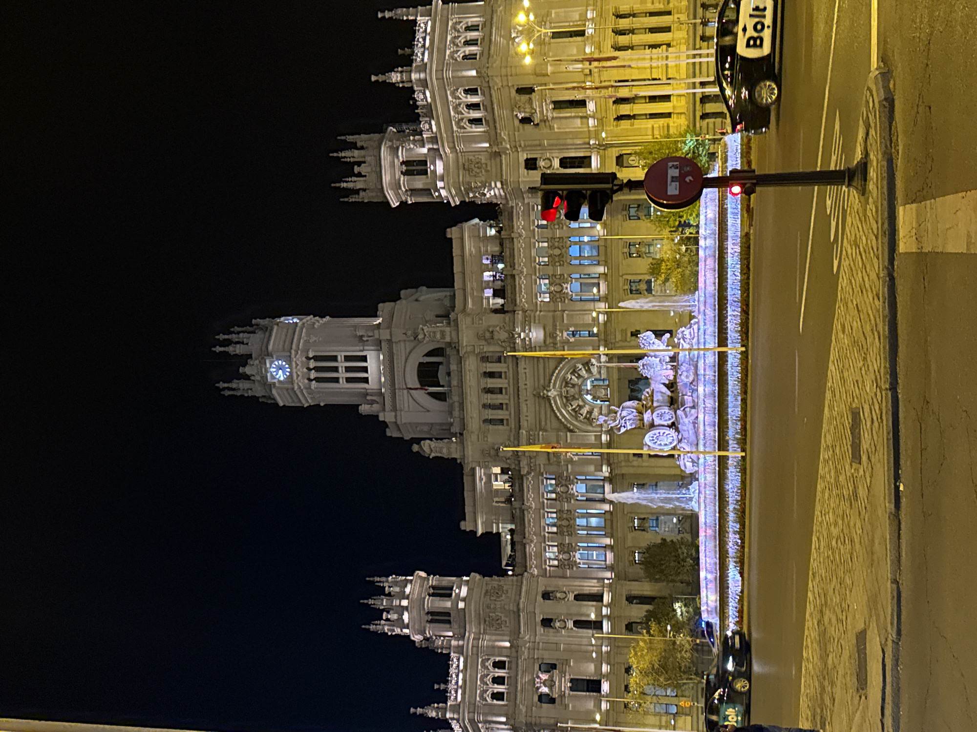 An eye-level, rotated shot shows a night view of the Cibeles Palace in Madrid, Spain. The Palace is centrally placed with its lit façade stretching from the bottom to the top of the frame. The building's clock tower and elaborate architectural details are highlighted in the night lighting.
In the foreground, the road and sidewalk are visible. A Bolt-branded car is parked near the curb. A traffic light post stands next to the road, displaying a red light. A "Do Not Enter" sign is atop the traffic light. A fountain with illuminated decorations stretches in the space between the road and the building. The background is the dark night sky.