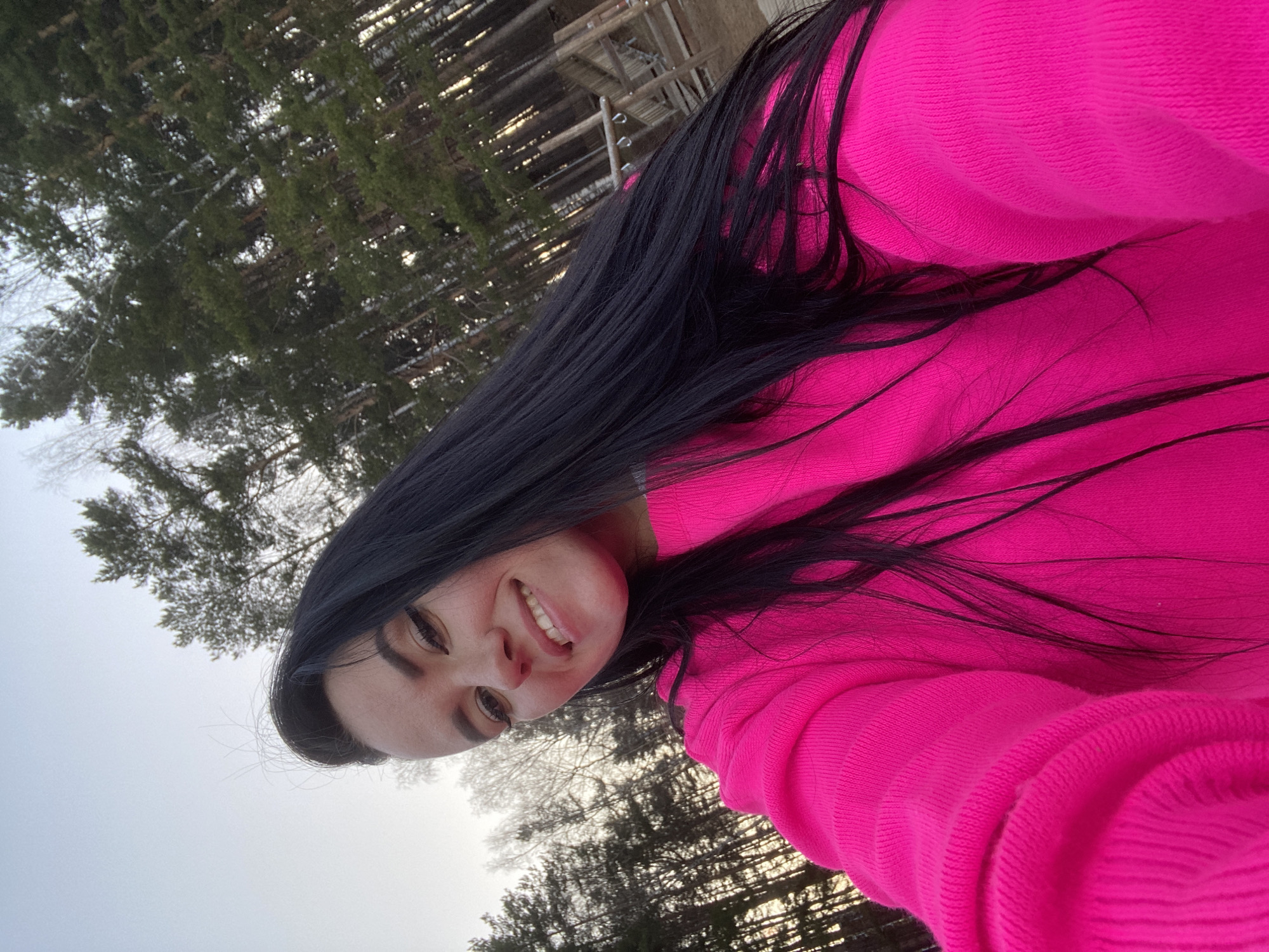 This close-up selfie features a young woman with long, dark hair, smiling broadly at the camera. She is wearing a vibrant, bright pink long-sleeved top or hoodie. Her face is angled slightly, with a cheerful expression and visible teeth.
The setting is outdoors, likely in Chaikovsky, Russia, characterized by a backdrop of coniferous trees, possibly pines or spruces, some of which appear to have a light covering of snow or frost on their branches. The sky is overcast and appears light grey, suggesting a cloudy day or soft, diffused light. In the upper background, through the trees, wooden structures resembling fences, scaffolding, or part of a larger outdoor facility are visible. The overall atmosphere suggests a cold, possibly wintery or early spring day. No text is visible in the image.