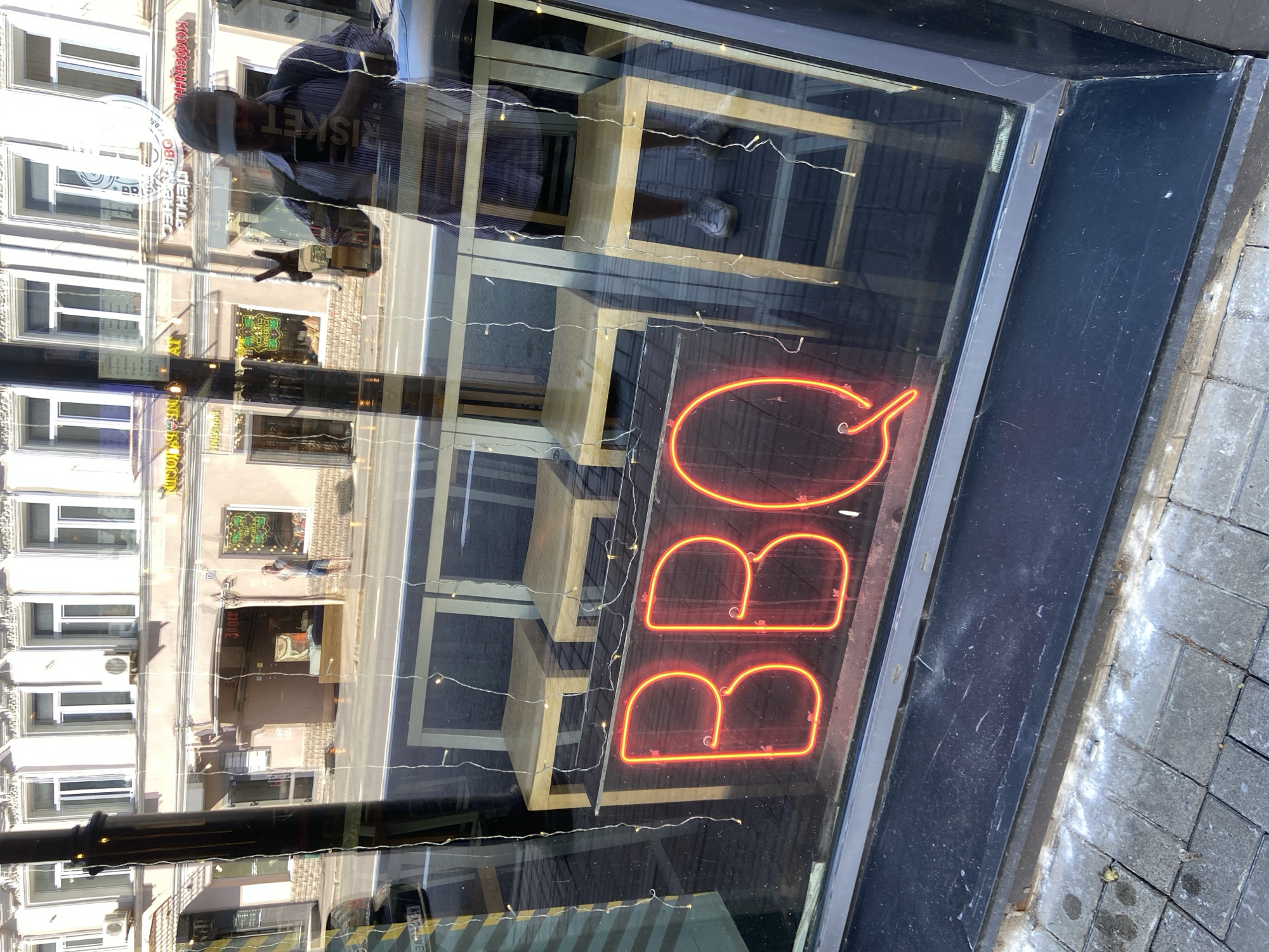 The image depicts a street scene reflected in a window. Inside the window, an illuminated "BBQ" sign is visible, suggesting a restaurant or eatery. Stools are stacked behind the sign. The reflection shows a street in what appears to be the city of Kazan, Russia, based on the presence of Cyrillic script on signs. A person is also reflected in the window, wearing a baseball cap, a light shirt, and holding a dark object that appears to be a smartphone. They are positioned in front of the storefront and are likely taking a photo of the "BBQ" sign. The street features multi-story buildings with windows and architectural details typical of urban areas. The foreground shows a segment of the sidewalk.