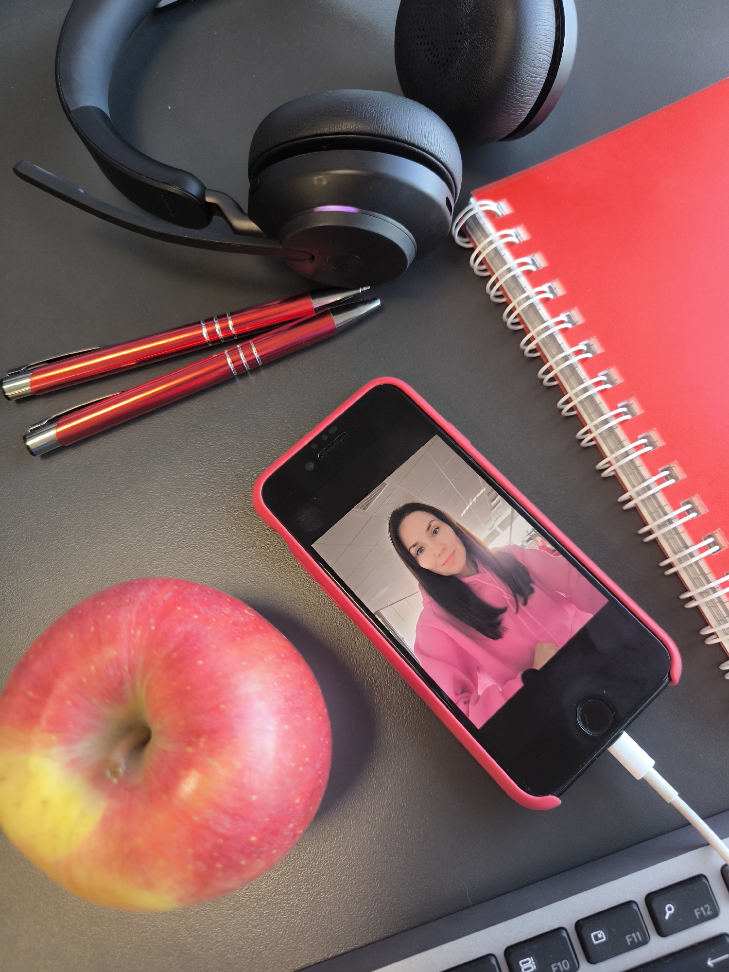 A still life photograph shows a desk scene. On a dark gray surface, a pair of black over-ear headphones lies near a red spiral-bound notebook. Two red pens rest adjacent to a red-cased black smartphone displaying a selfie of a woman wearing a pink hoodie. A partially yellow apple sits to the left of the phone. A white charging cable is plugged into the bottom of the phone, extending toward a gray and black keyboard at the bottom of the frame. The location is unidentified, with no clear indication of Riga, Latvia.