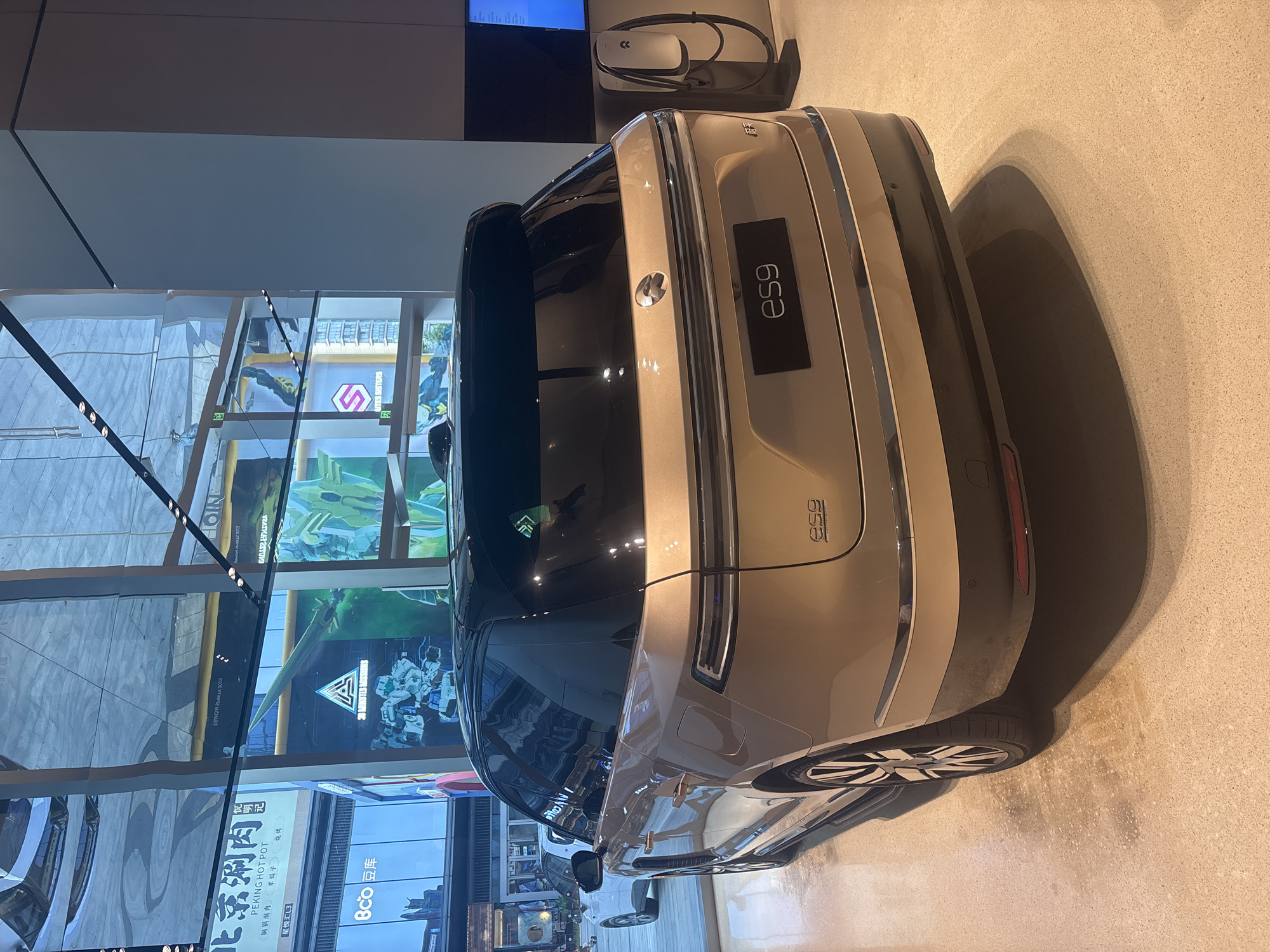 The image captures the rear three-quarter view of a gold NIO ES8 electric SUV parked indoors, likely in a showroom. The vehicle is positioned on a speckled beige floor. Behind the car, a large window reflects the environment, showing various signs and advertisements. To the right of the car, a white NIO electric vehicle charging station is visible. The car's branding, including the NIO logo and "ES8" badging, is clearly discernible. The overall scene suggests a retail or exhibition space, consistent with a car dealership in a modern urban setting such as the Suzhou Industrial Park, China, indicated by the reflections of Chinese text and logos in the background.