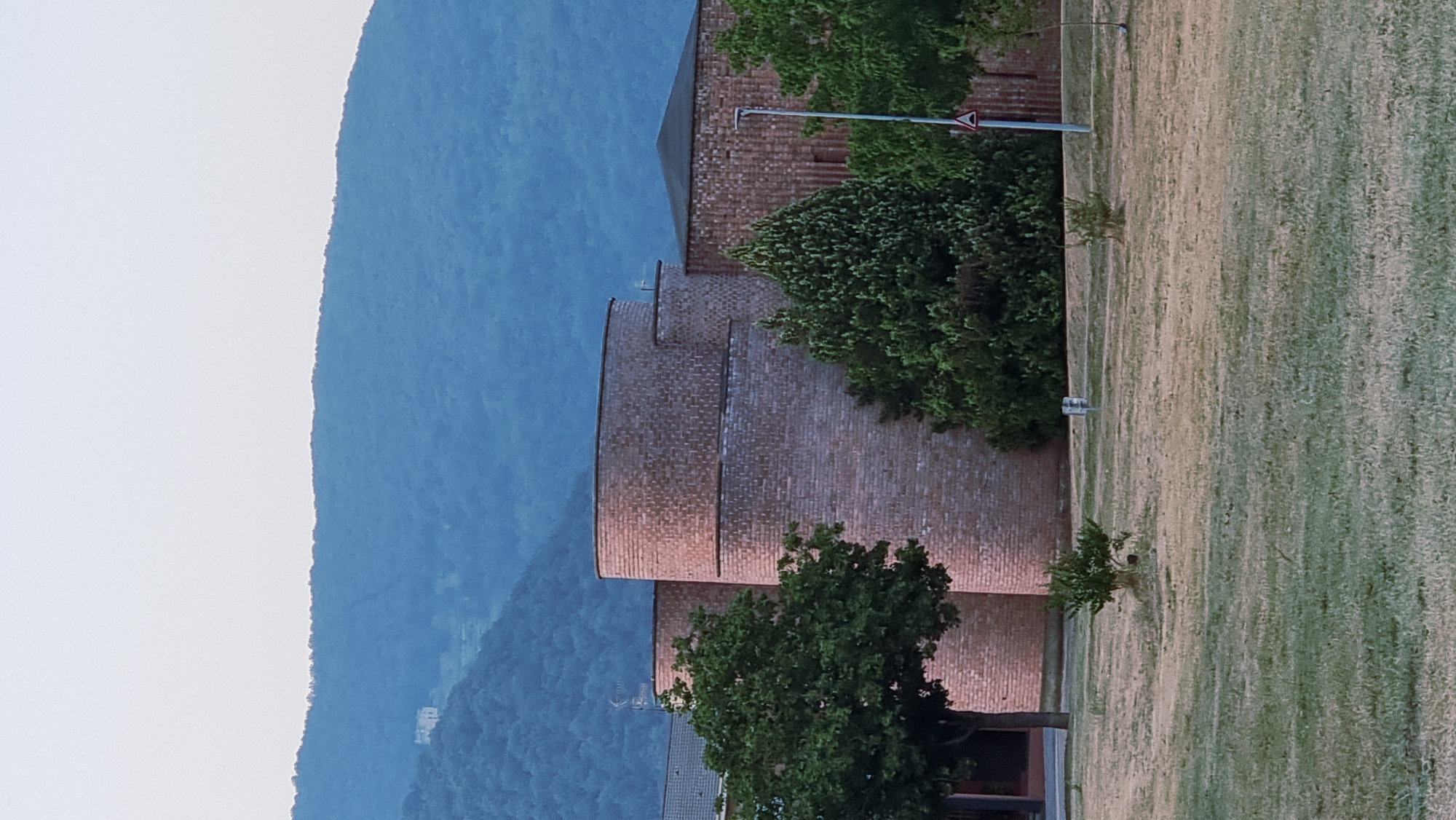 The image shows a building with rounded brick towers, set against a backdrop of blue-green forested hills. The sky is a pale, hazy blue. In the foreground, a dry, mown lawn has scattered patches of green grass. A few small trees and bushes dot the lawn. The building's brickwork is detailed, with visible mortar lines. To the right of the building, a tall pole with a traffic sign is visible. The overall scene suggests an exterior view of a modern structure in a semi-rural or park-like setting. A small, light-colored building can be seen on a distant hillside.