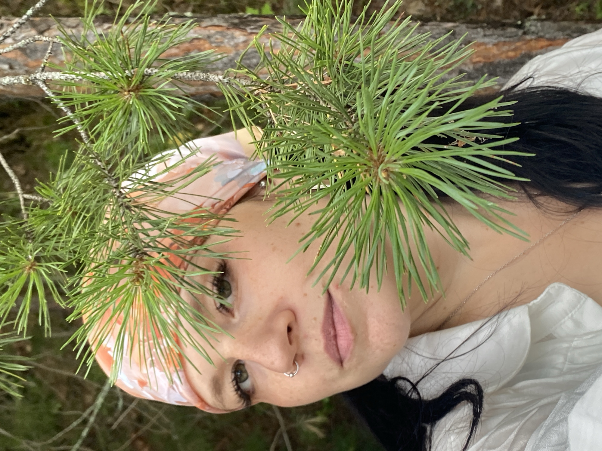 The image is a close-up portrait of a woman partially obscured by pine branches. She is wearing a light-colored bandana with a pink and orange pattern and a white shirt. She has a silver hoop nose ring and a thin silver necklace. Her hair is dark and long. The background appears to be a forest, with a pine tree trunk visible on the right. The lighting suggests it is daytime. The image was taken in the city of Chaykovsky, Russia.