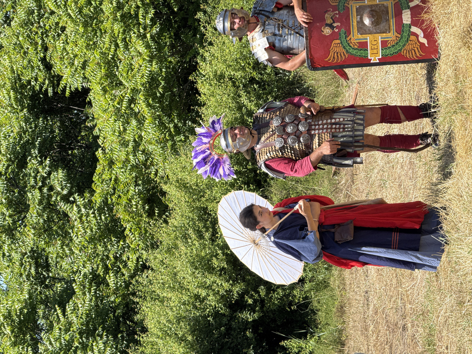This image, captured in Gigen, Bulgaria, depicts three individuals in historical Roman-style costumes standing outdoors in a sunlit field. On the left, a woman with dark hair wears a layered blue and light blue tunic, accented by a red sash draped over her left shoulder and arm. She holds a white parasol with a wooden handle over her right shoulder. Adjacent to her, a man portraying a Roman centurion wears a metallic helmet adorned with a prominent purple feathered crest, a red long-sleeved tunic, and elaborate bronze-colored segmented armor (lorica segmentata) embellished with multiple circular medallions. He holds a dark stick or staff in his right hand. To the right, a second man in Roman soldier attire wears a metallic helmet, segmented armor, and a red undergarment. He holds a red rectangular Roman scutum (shield) upright, resting on the ground, which features a golden wreath, a lion motif, and the inscription "LEG" alongside a central metallic boss. The foreground is composed of dry, golden-brown grass with scattered green weeds, while the background consists of dense, verdant foliage, including trees and bushes, under a clear blue sky.