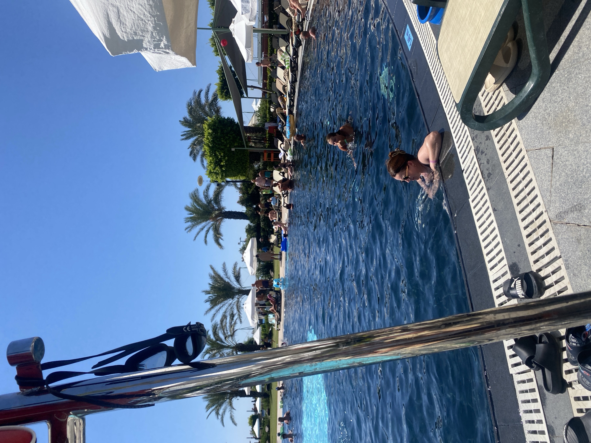 An outdoor swimming pool scene is depicted on a sunny day, likely at a resort in Aksu, Turkey. In the foreground, a chrome railing with black swimming goggles hanging from it is partially visible on the left. The pool itself is dark blue and has several people swimming or relaxing in it. To the right of the pool, lounge chairs are arranged, with some occupied. Palm trees and white umbrellas are visible in the background, suggesting a tropical or Mediterranean setting. The sky is a clear, bright blue. Some beach towels and flip-flops are scattered around the pool area.