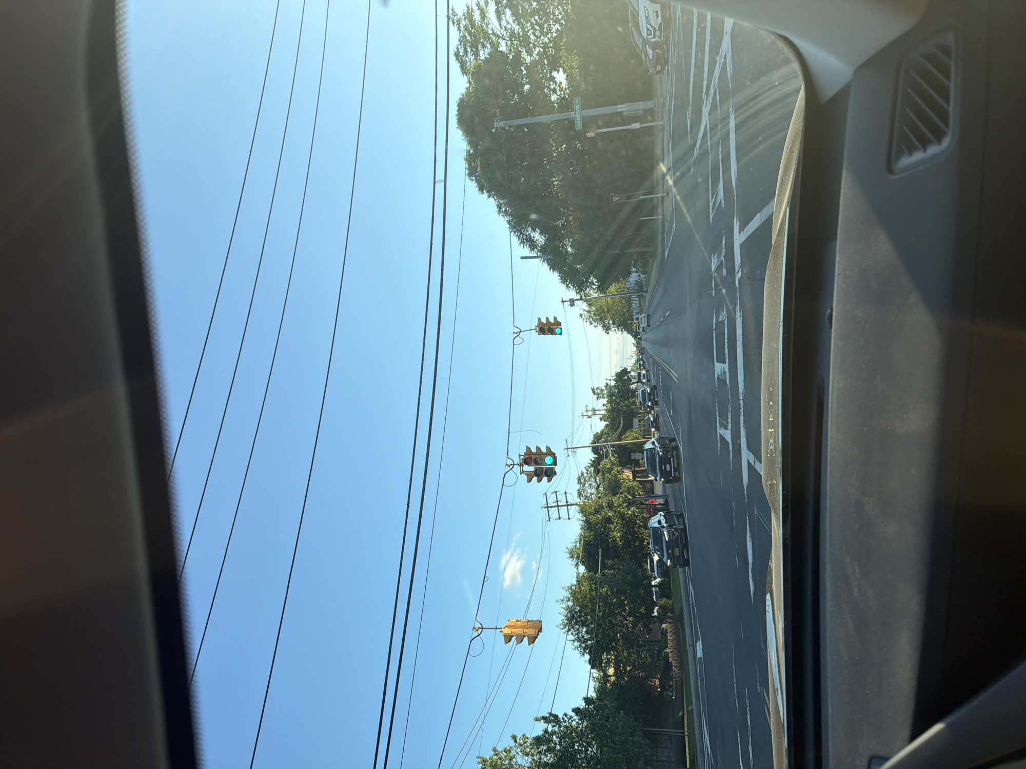 This is a point-of-view shot from a vehicle driving on Harrison Avenue in Riverhead, NY, USA. The sun is shining, and the sky is clear blue, with several power lines crisscrossing overhead. The traffic lights are green, indicating that it is safe to proceed. Several cars are driving on the road, and there are trees and buildings on either side. The road is marked with white lines, and there are crosswalks. The image appears to have been taken during the day, possibly late afternoon due to the angle of the sun.