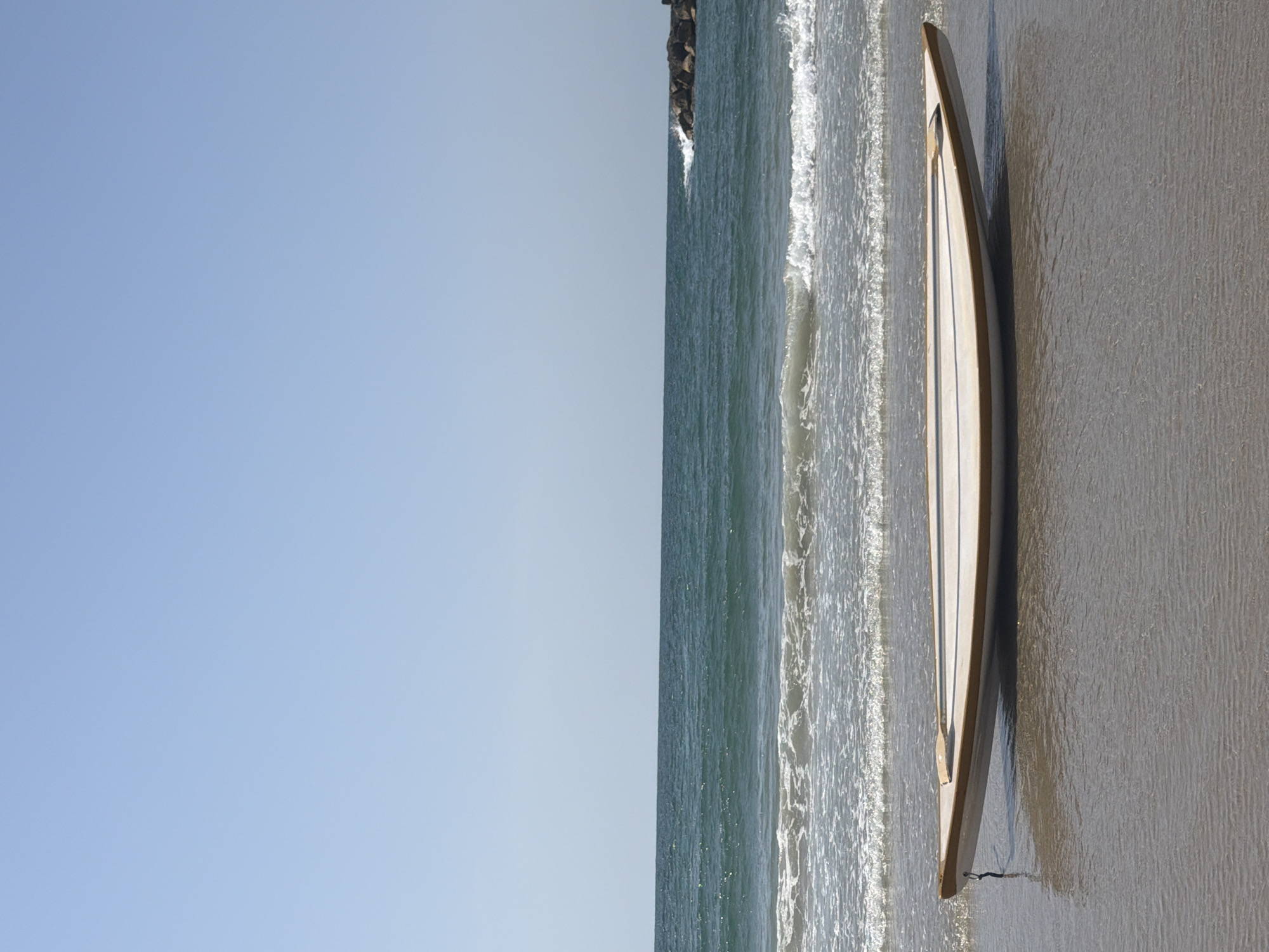 The image depicts a surfboard lying on the sand at a beach in Tel-Aviv, Israel. The sky is clear and blue. The beach is partially covered by water with small waves. A dark rock pier is visible in the distance.
