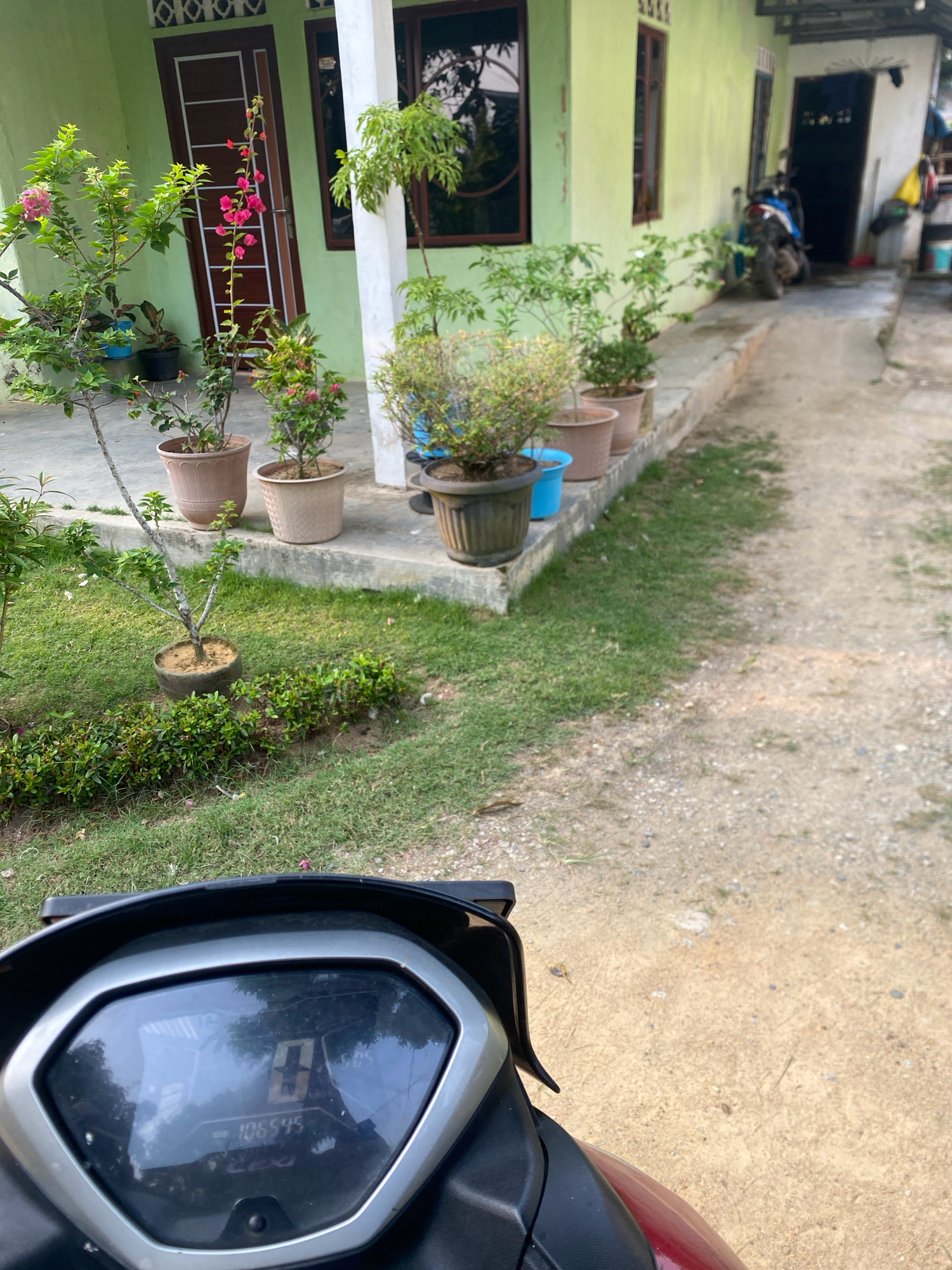 The image shows a view from a motorcycle, looking towards a light green house with a white pillar and brown trim around the windows and doors. Several potted plants, including flowering bougainvillea and other greenery, are arranged on the concrete path and steps leading to the house. A blue motorcycle is parked on a ramp to the right of the entrance. The foreground shows the dashboard of the motorcycle with a digital display showing "0" and "106545". The ground is a mix of dirt and gravel. The scene appears to be in a residential area, possibly in Batam, Indonesia, given the architecture and vegetation.