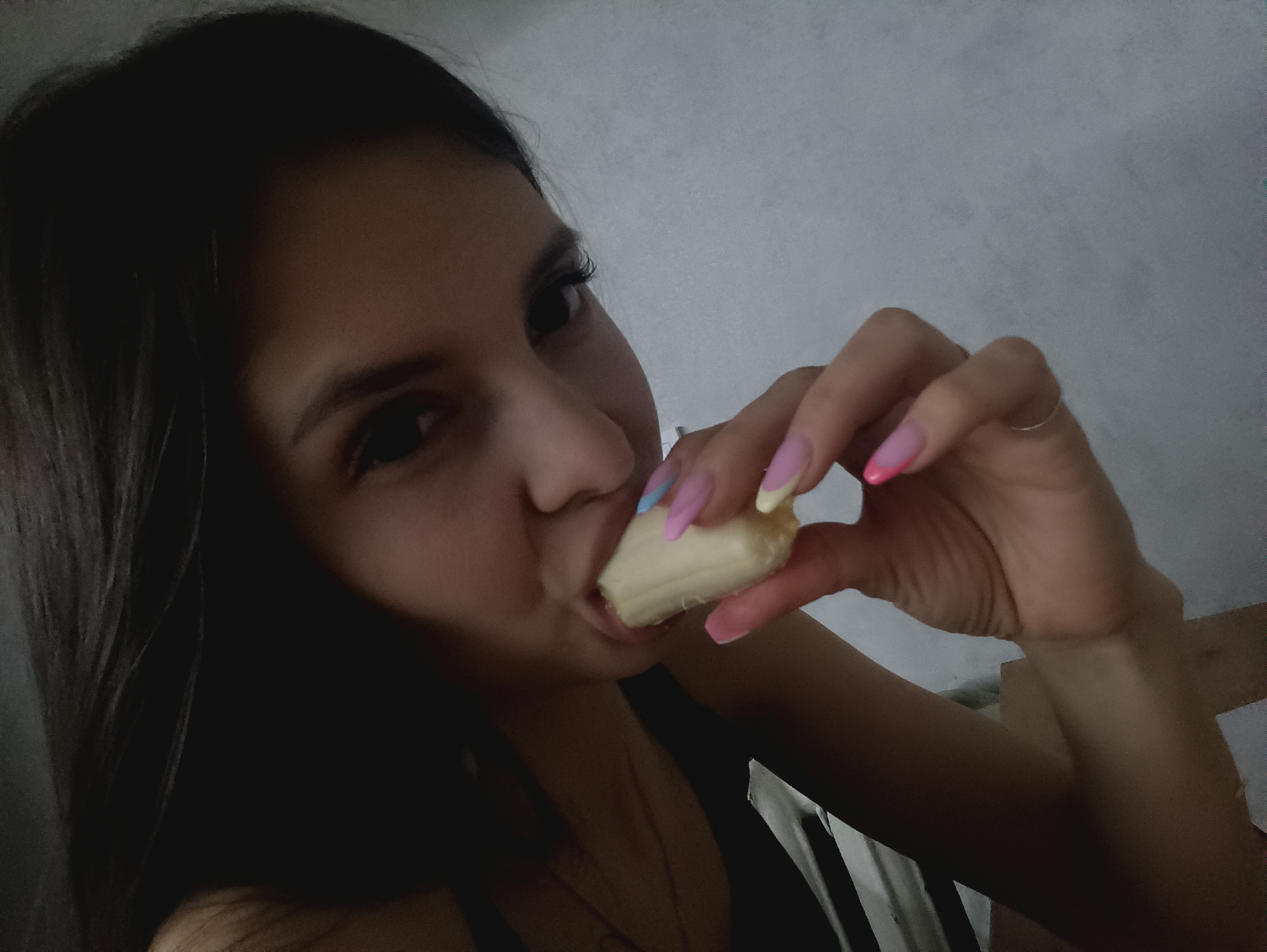 A woman with long, dark hair is holding a peeled banana to her mouth. Her fingernails are long and painted with pastel colors. She is wearing a black top. The background is a plain, light gray wall. The location is Unknown, Unknown.