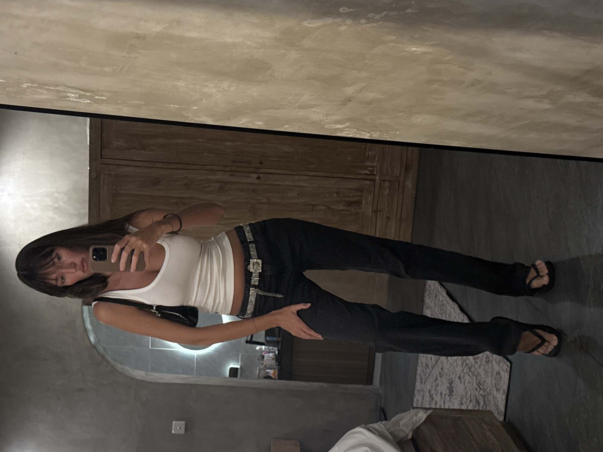 A full-body, waist-up shot captures a woman from the chest up, holding a smartphone and taking a mirror selfie. She is wearing a white tank top, black jeans, a sparkling silver belt, and black flip-flops. The background consists of a textured gray wall, a wooden wardrobe, and a dimly lit room. The overall aesthetic suggests a modern, urban setting, possibly in a hotel room or apartment. The image appears to have been taken indoors, with soft lighting that casts subtle shadows. The woman's expression is neutral, and her posture is relaxed. The scene is devoid of any other people or significant activity. The presence of a smartphone indicates the act of self-documentation.