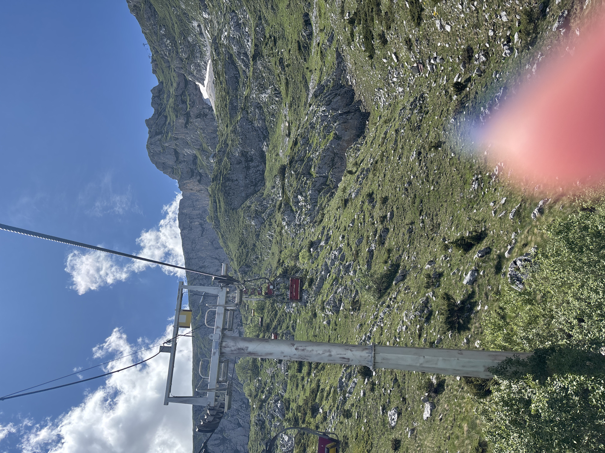 This image shows a ski lift ascending a steep, rocky, and grassy mountainside under a bright blue sky with scattered clouds. The ski lift is composed of several red cabins suspended from a steel cable, supported by a tall metal pylon in the foreground. The mountain slope is covered in green grass, scattered rocks, and some sparse bushes and trees. In the distance, the mountain peaks are rugged and rocky, with patches of snow visible on some higher elevations. The image appears to be taken from a moving ski lift, looking upwards along the slope. The location is specified as Pašina voda, Montenegro. There are no people visible in the image. A reddish-pink lens flare is present in the lower right quadrant of the image, obscuring some of the mountain slope.