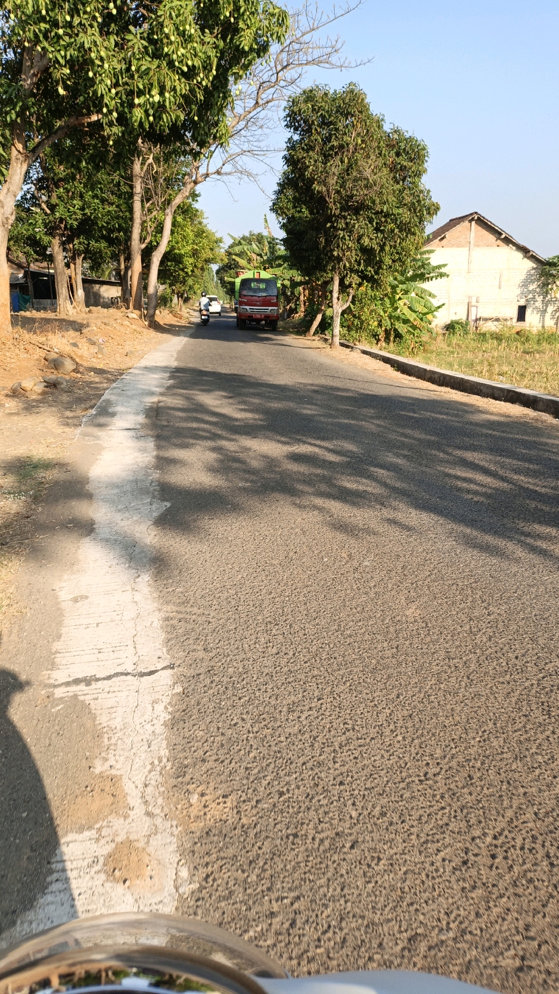 The image depicts a road scene in Kuwasen, Indonesia. In the foreground, the perspective is from a moving vehicle, likely a motorcycle, showing the asphalt road and a portion of the vehicle's chrome handlebar. The road is lined with trees on both sides. On the left, there are trees with fruit, and the ground is rocky and dry. On the right, there are more trees and a building with a yellow facade. A red truck is visible driving on the road, along with a white car and a motorcycle. The sky is clear and blue, indicating a sunny day. The overall environment suggests a rural or semi-rural area within Kuwasen.