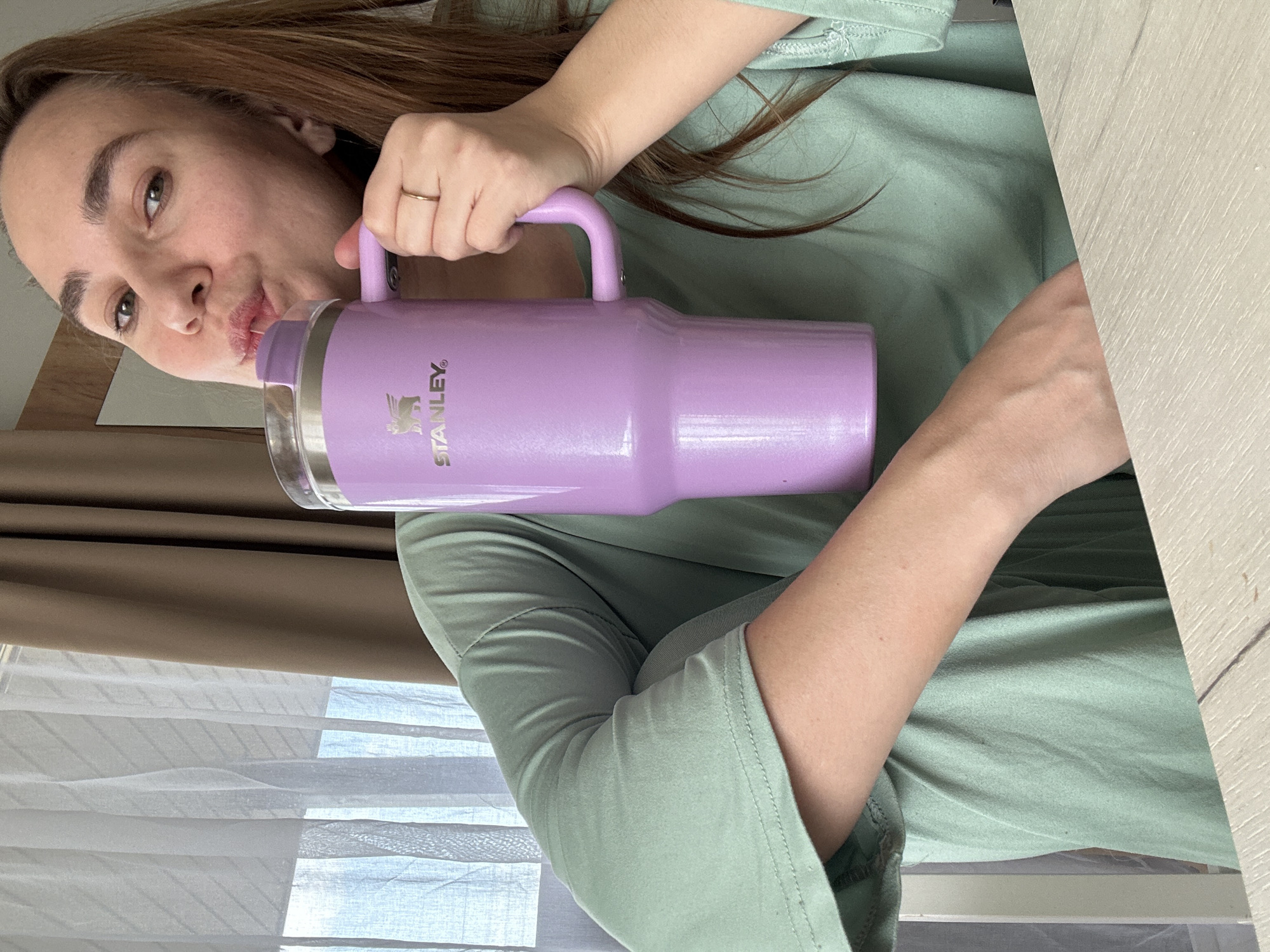 A young woman is depicted in a close-up, angled portrait, holding and appearing to drink from a large, light purple Stanley tumbler. Her lips are pursed around a transparent straw that extends from the tumbler's lid. She has long, brown hair and is wearing a simple, light green top. A gold-colored ring adorns her right ring finger, while she holds the tumbler by its handle with her left hand. The "STANLEY" brand name and its distinctive bear logo are prominently visible on the side of the tumbler.
The setting appears to be indoors, with natural light suggesting it is daytime. In the background, white window blinds are visible, partially covered by a brown curtain or frame, indicating a window. To the right, a light-colored wooden surface, possibly a table or desk, is present. The woman looks directly at the camera, conveying a casual and engaged expression. The location context for this image is Kharnavy, Russia.