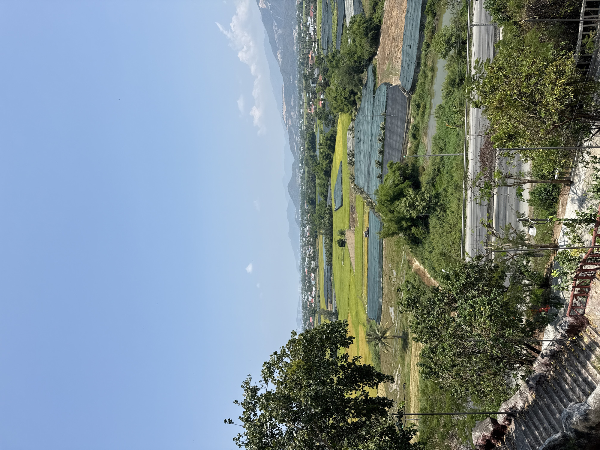 The image depicts a landscape in Bắc Nha Trang, Vietnam. The sky is a clear blue with some light clouds. A mountain range is visible in the distance. In the midground, there are fields with a variety of crops, including rice paddies and areas covered by netting. A road runs parallel to the fields, bordered by trees and other vegetation. In the foreground, there are trees and a stone structure, possibly part of a staircase or wall. The scene appears to be taken from an elevated vantage point.