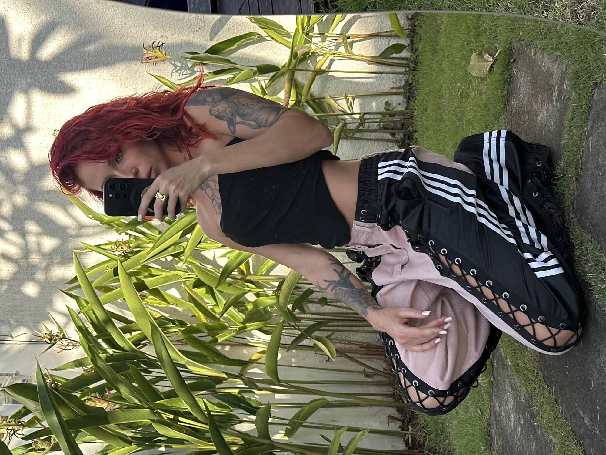 A woman with bright red, wavy hair is kneeling in front of a wall with a white textured finish, partially covered by tall green plants. She is taking a selfie with a black smartphone held in her right hand, which is positioned near her face. She is wearing a black strapless top and a pair of pink and black pants with lace-up details on the sides and white stripes on the black sections. The pants have an athletic style. Tattoos are visible on her left arm. The ground is covered in grass and a concrete path. The scene appears to be outdoors, likely in a garden or courtyard, given the plants and natural lighting. The sunlight casts shadows from the plants onto the wall. The woman is looking at her phone screen. The overall impression is a casual, outdoor portrait. The location is identified as North Kuta, Indonesia.