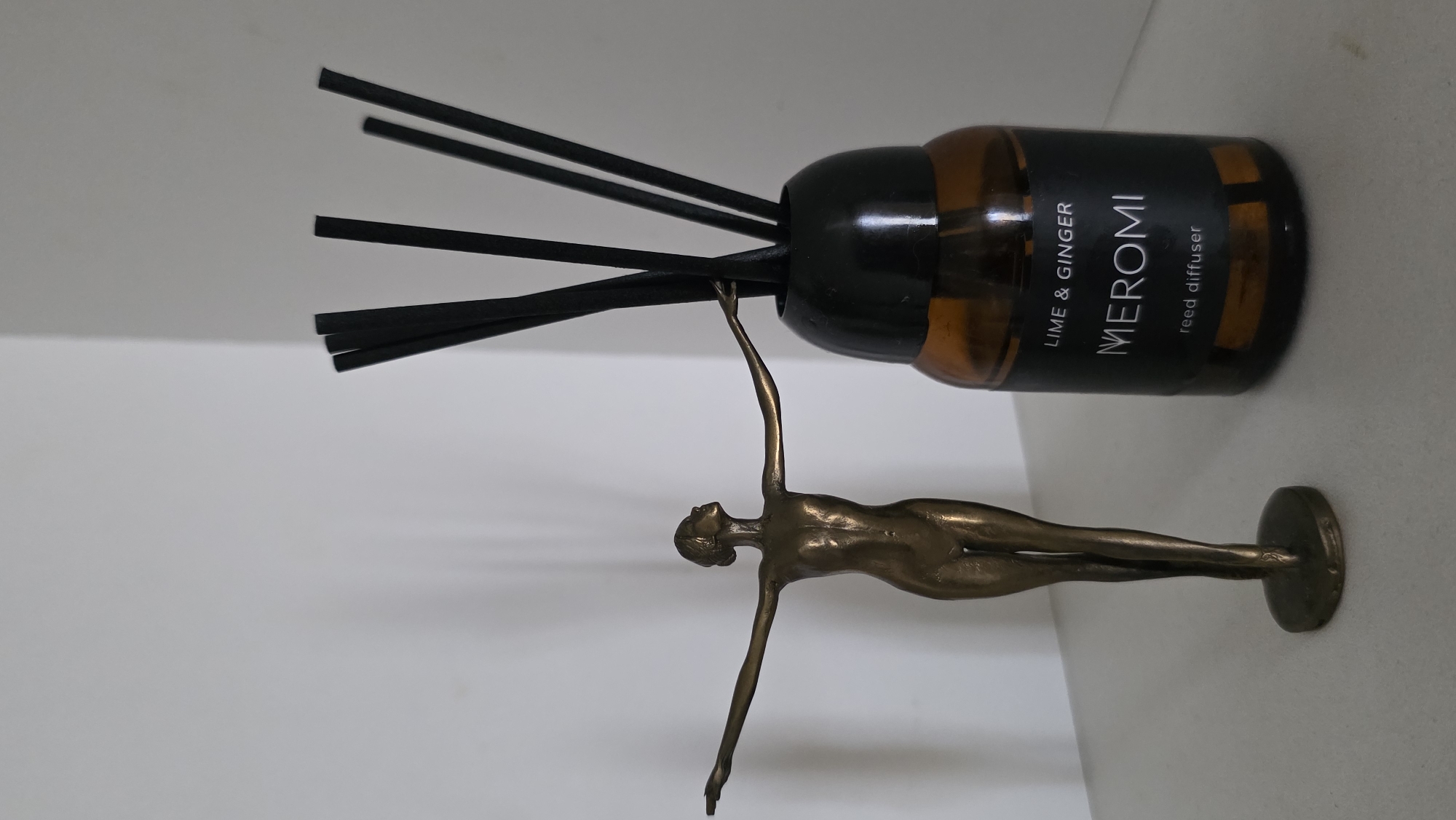A brass figurine of a nude woman with outstretched arms stands to the left of a glass reed diffuser bottle. The figurine is on a circular base. The reed diffuser has a dark brown glass bottle with a black cap and black reeds inserted into it. A black label on the bottle reads "LIME & GINGER" at the top, "MEROMI" in the center, and "reed diffuser" at the bottom. The objects are placed on a light-colored surface against a plain white background. The scene is lit from the front, casting shadows behind the objects. The overall lighting is soft. The image does not contain any information about the location in Tambov, Russia.
