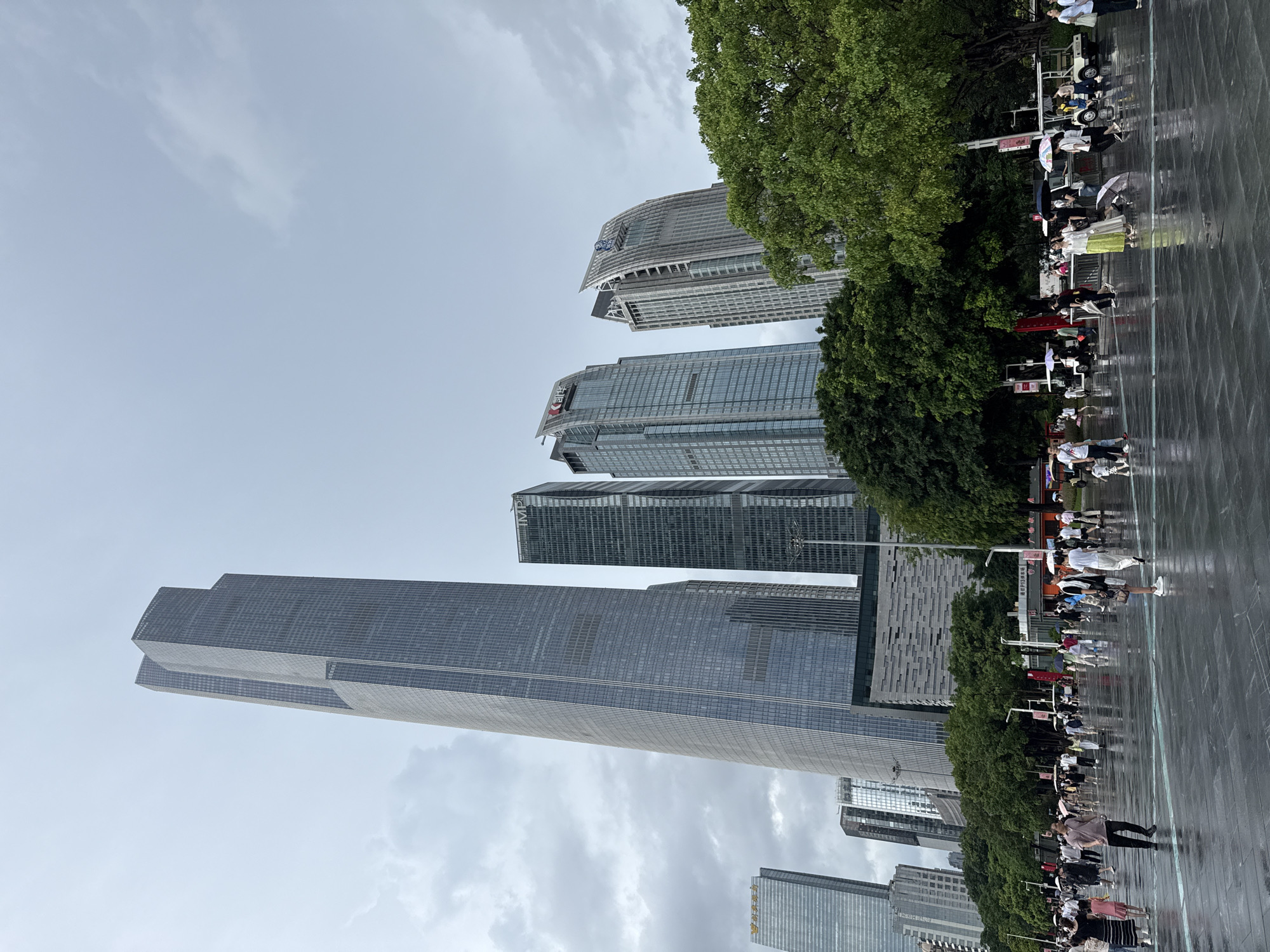 The image depicts an outdoor scene in Tianhe District, China, likely a public square or plaza, with a prominent skyline of modern skyscrapers in the background. The ground is wet and reflective, suggesting recent rainfall, and several people are walking around, many holding umbrellas, indicating a damp or rainy day. The architecture is characterized by tall, glass-facade buildings, some with distinctive angular tops. Lush green trees are visible in the middle ground, framing the urban landscape. A variety of people are scattered across the open area, dressed in casual summer clothing. Some individuals are walking, while others appear to be standing or gathering. Small carts or vehicles, possibly for transportation or maintenance, are parked in the vicinity. The sky is overcast with clouds.
