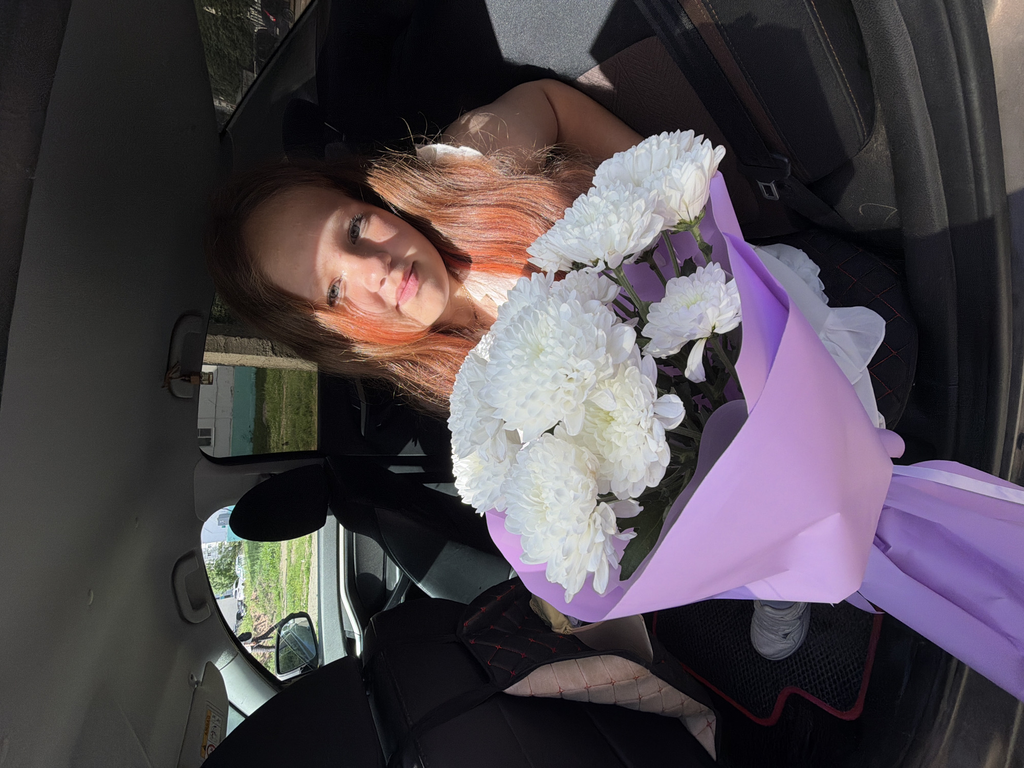 A young girl with long, reddish-brown hair sits in the back seat of a car, holding a bouquet of white chrysanthemums wrapped in purple paper. She is looking towards the camera with a slight smile. Sunlight is streaming into the car, casting shadows and highlighting her hair. The interior of the car is visible, including the seats, a seatbelt, and a car mat on the floor. Outside the car, a residential area in Chaikovsky, Russia, can be seen through the windows, with buildings and trees.