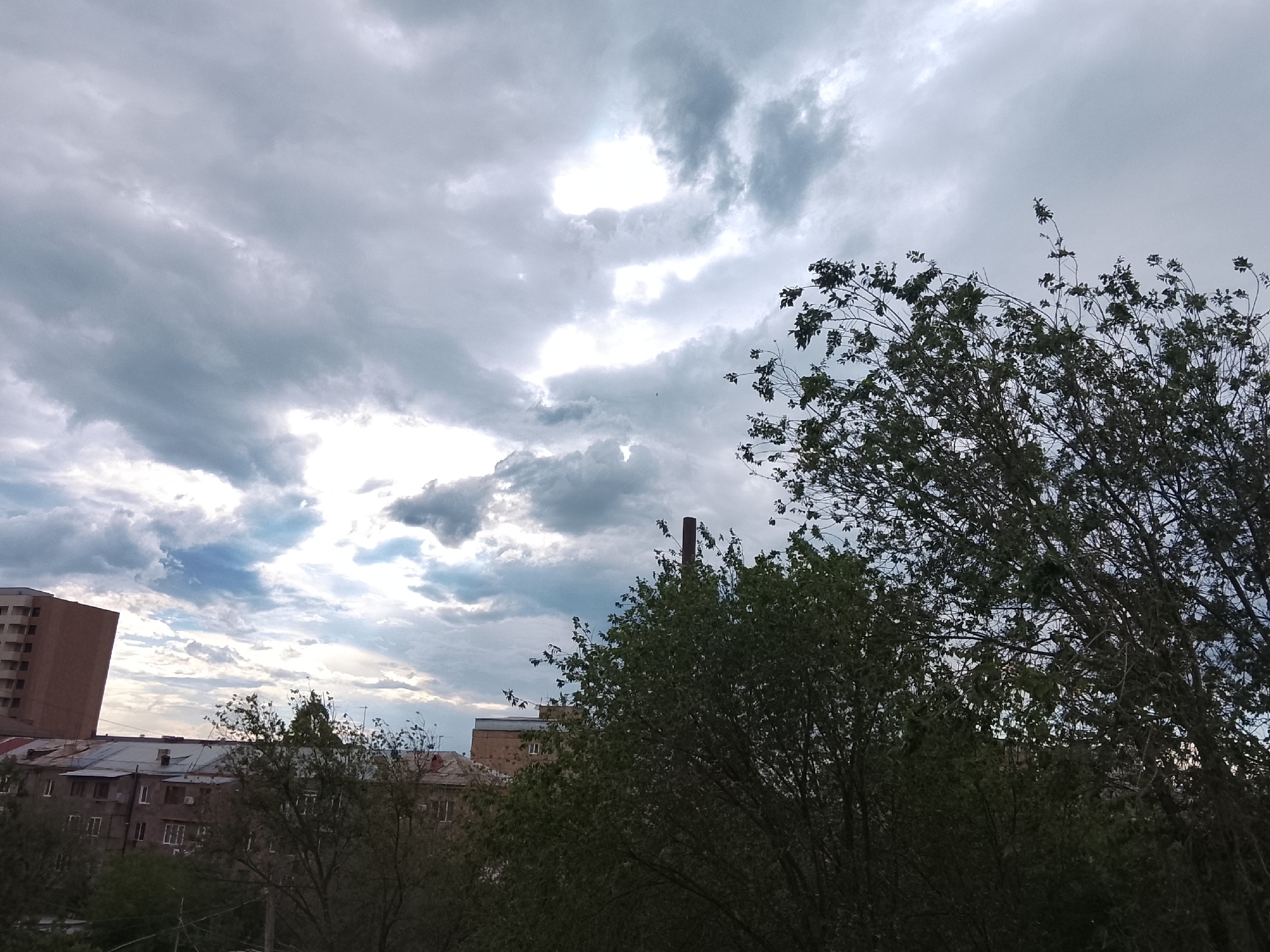The image captures an urban or suburban landscape dominated by a dynamic, cloudy sky. The sky features a mix of dark grey and bright white clouds, with patches where sunlight appears to break through, illuminating certain areas.
In the foreground and mid-ground, dense green trees with numerous leaves and branches fill a significant portion of the frame, especially on the right side and extending across the bottom. The trees obscure parts of the background, creating a layered depth.
Behind the trees, several buildings are visible. On the far left, a tall, multi-story building with a light brown facade stands prominently. Further into the background, other lower-rise buildings with dark roofs and multiple windows can be seen, suggesting residential or commercial structures. A distinct dark, chimney-like structure rises above the trees in the center-right mid-ground.
The time of day appears to be daytime, characterized by the strong light penetrating the clouds, hinting at either a clearing after rain or an impending change in weather. The overall impression is one of an overcast or partly cloudy day. There is no discernible text in the image.