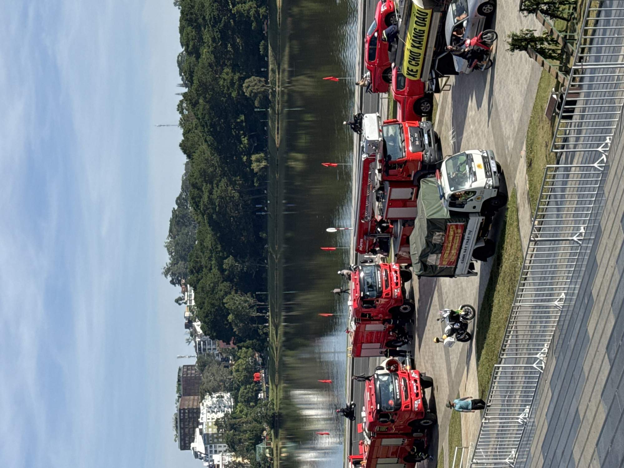 The image depicts a sunny day in Da Lat, Vietnam, likely during an event or parade. The primary subjects are several red fire trucks and a white utility truck parked in a row along a waterfront promenade. A few people are visible, some near the vehicles and others standing or walking on the promenade. One person is seen on a motorcycle.
The setting is a picturesque lakeside, with lush green trees and a cityscape featuring buildings in the background. The calm water reflects the trees and the sky. The promenade appears paved with gray tiles and lined with a metal railing. Red flags are strategically placed along the edge of the promenade.
The overall atmosphere appears calm and organized, suggesting an official gathering or demonstration. The bright sunlight indicates it is daytime, likely mid-morning or afternoon. The presence of fire trucks and the organized display suggest a civic event or perhaps a training exercise.
Visible text on a yellow banner attached to one of the trucks reads "KE CHO KANG DÀU". Another truck has text that is partially obscured but appears to be "CẢNH SÁT TRẬT TỰ".
Notable details include the vibrant red of the fire trucks, the clean lines of the modern promenade, and the reflection in the water, all contributing to a visually appealing scene. The clear blue sky with faint clouds adds to the pleasant weather conditions.