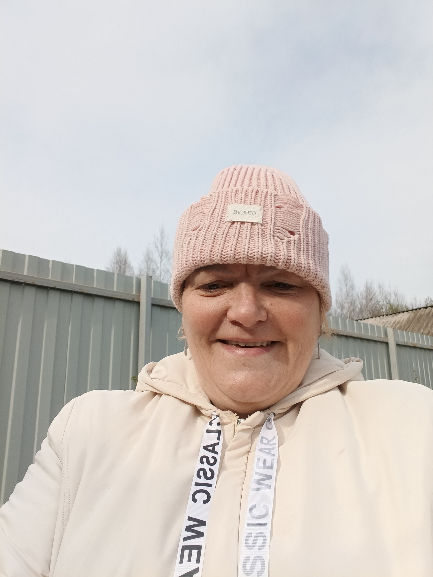 A smiling woman is captured in a close-up, outdoor selfie or portrait, likely in Красный Маяк, Russia. She appears to be middle-aged or older, with a cheerful expression, looking directly at the camera. She is wearing a light pink knitted beanie hat that features a distressed, ripped appearance and a small white label. The text on the label, when inverted, reads "GENIOUS". Her attire also includes a light beige or off-white hooded jacket. The jacket's drawstrings prominently display repeated text, which when inverted, reads "CLASSIC WEAR".
The background features a light grey corrugated metal fence, indicating an outdoor setting. Behind the fence, sparse, bare trees are visible, silhouetted against a bright, clear, light blue sky. The sunny weather and lack of leaves on the trees suggest either early spring or late autumn. The overall impression is one of a relaxed, casual moment in an open, possibly semi-rural environment.