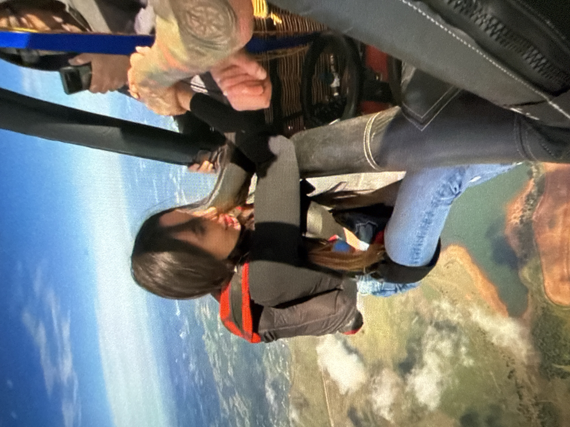 A woman is mid-air during a tandem skydiving experience. She wears a black jumpsuit with red detailing, light blue jeans, and a black harness. An instructor is attached to her, visible by tattooed arms holding onto the harness. The background depicts a view of land with green fields, bodies of water, and blue skies. The location is Unknown, Unknown.