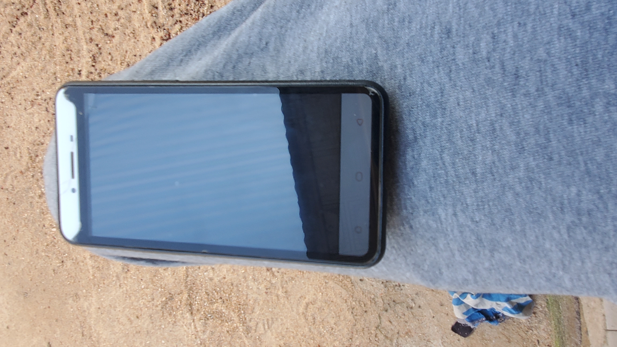 A smartphone is resting on a person's lap, on a surface that appears to be sand or loose soil. The smartphone is black with a white bezel and a reflective screen that shows a distorted image of what is likely the sky and a dark, wavy horizon. The phone is positioned vertically. The person's lap is covered with a light blue-grey fabric, possibly sweatpants. In the lower left corner of the image, a piece of fabric with blue and white stripes is partially visible, along with a small black object. The background consists of a light brown, sandy area with scattered small pebbles. This scene is set outdoors, possibly in Jalingo, Nigeria, given the sandy environment, which is common in parts of the city.