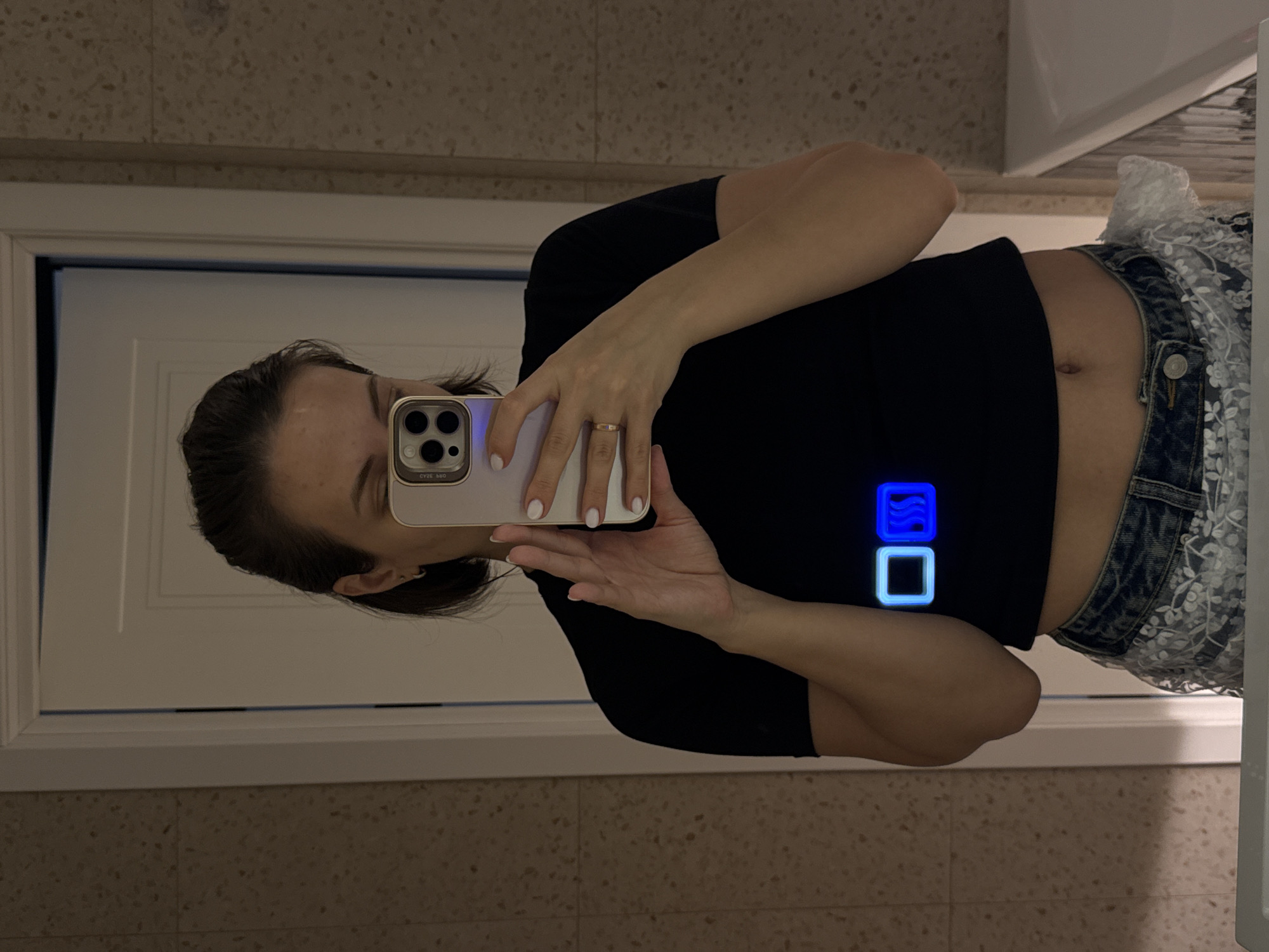 An image, rotated 90 degrees clockwise, captures a person taking a mirror selfie indoors. The primary subject is a young adult, likely female, with dark hair pulled back, seen from the waist up. She is wearing a black, short-sleeved crop top that exposes her midriff and belly button, paired with denim shorts featuring a white lace overlay. A simple gold-colored ring adorns her right ring finger, and her fingernails are painted white.
She holds a light-colored smartphone, possibly an iPhone given the distinctive camera array, in her right hand to capture her reflection. Reflected text, "СУЭ 54D" (appearing reversed due to the mirror), is visible on the phone's screen. On the black crop top, two digitally added or reflected glowing blue square icons are visible near her right elbow: one solid blue with wavy lines, and the other a hollow outline.
The setting appears to be a bathroom or a similar indoor space with a large mirror. The background, visible in the reflection, consists of light beige/tannish speckled tiles on the wall, suggesting a durable surface. A white door frame or cabinet is visible on the left side of the reflection, and a white sink or counter edge can be seen on the right. The lighting is artificial and somewhat dim, casting a warm glow. The overall scene depicts a casual moment of self-photography in a private setting in Pervomaysky, Russia.