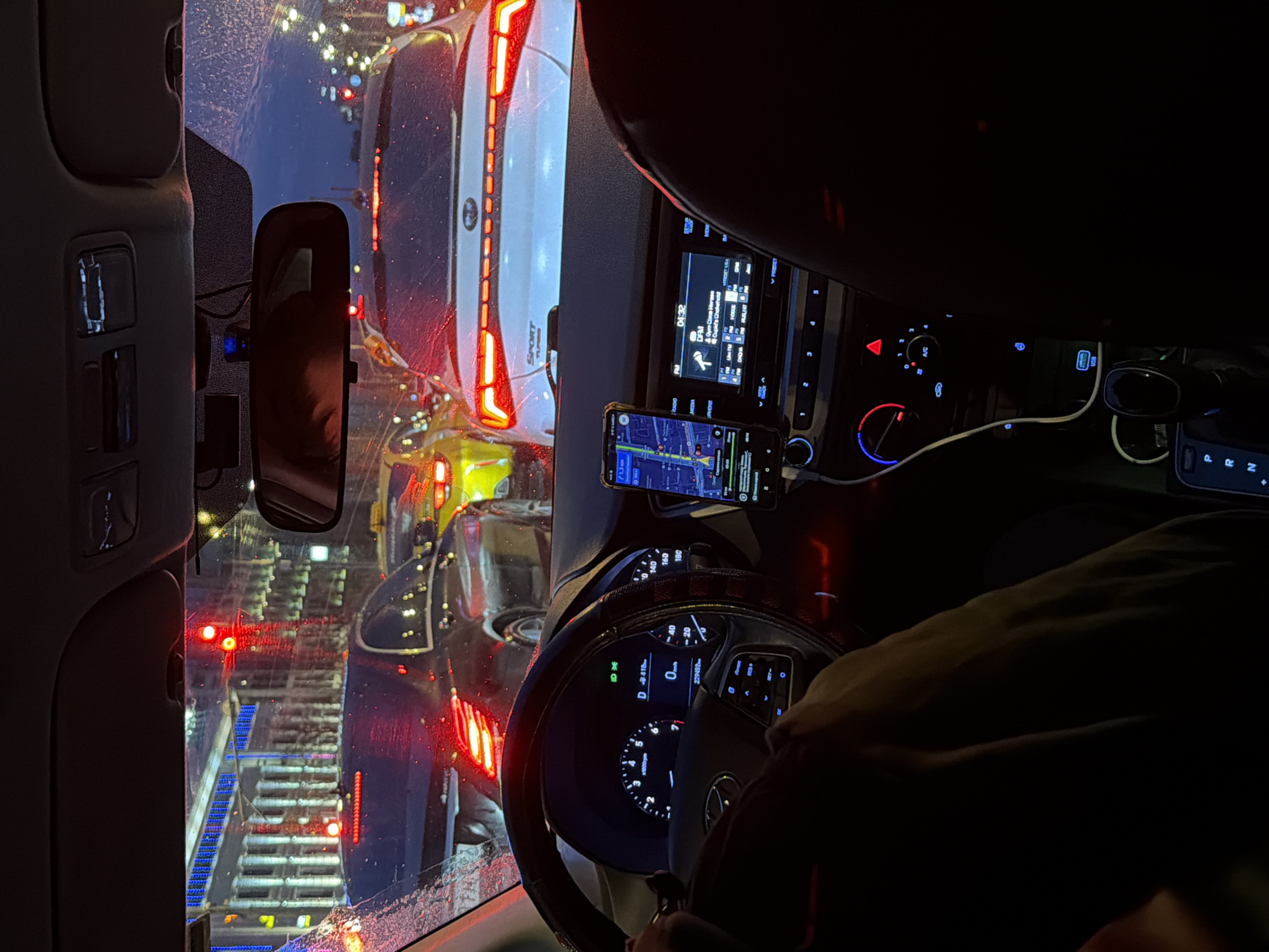 The image captures a view from inside a car, likely during heavy traffic at night. The driver's hand is visible on the steering wheel. A navigation app runs on a smartphone mounted on the dashboard. The instrument panel displays a speed of 0 km/h, indicating the car is stationary. Outside, other vehicles are visible, with red taillights shining, suggesting a traffic jam on a wet road. The car in front is a white Hyundai Tucson Sport. Through the windshield, the lights and structure suggest an elevated tram/train line or overpass, potentially indicating the image was taken in a densely populated area in Moscow, Russia.