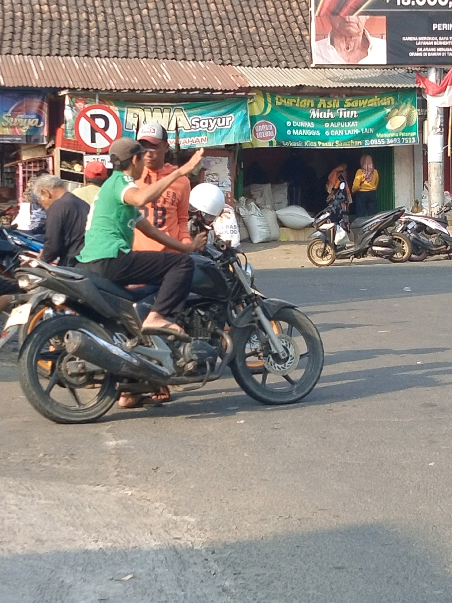The image captures a street scene in Sawahan, Indonesia, with a focus on a group of people and motorcycles in front of a market or shop. In the foreground, a black motorcycle is parked, with its front wheel and part of the engine visible. A man in a green shirt and black pants is seated on a black motorcycle, gesturing with his right hand. Another man in an orange shirt is standing next to him, also gesturing with his right hand. A person wearing a white helmet is partially visible behind them. Behind these individuals, more motorcycles are parked, and people are seen in the background, some entering or exiting a building with a green awning. The building's awning displays text in Indonesian, including "Durian Asli Sawahan Mak Tun" and "Nomor Telepon 0855 3693 7513," indicating a business selling durian in the Sawahan market area. The environment appears to be a bustling street, likely in a commercial district, with the sun casting shadows on the asphalt road. A "No Parking" sign with a red circle and diagonal line over the letter "R" is also visible on the left side of the image. The overall scene depicts a lively street activity with vendors and customers interacting in the Indonesian context of Sawahan.