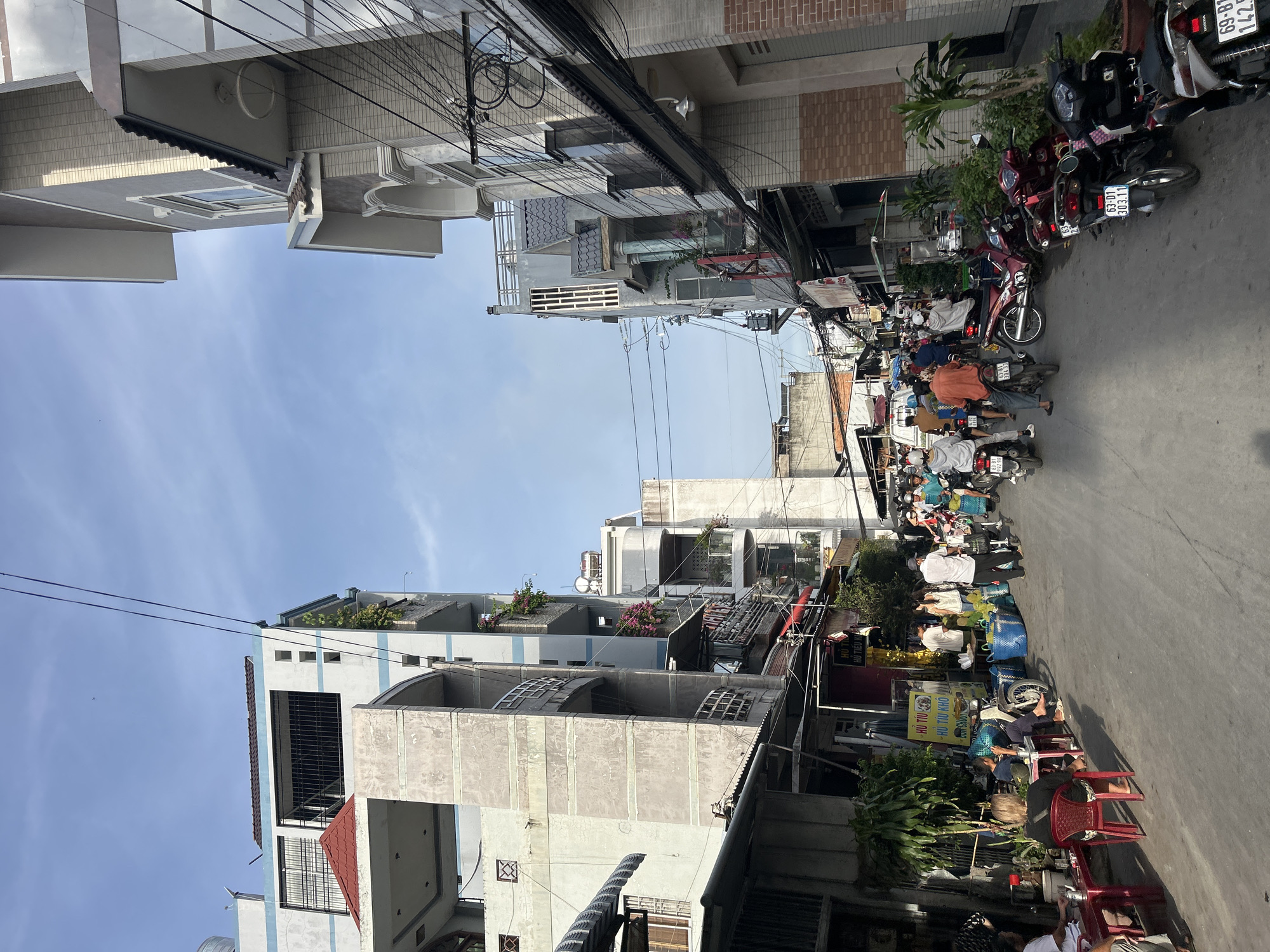 This is a ground-level, eye-level, full-shot photograph of a street scene in An Hữu, Vietnam. The street is paved and appears to be narrow, with two-story and multi-story buildings lining both sides. The buildings are a mix of modern and older architecture, with balconies and awnings visible. There is a significant amount of electrical wiring strung across the street and between buildings.
The street is bustling with activity. Several people are on foot, some walking and some gathered around stalls. There are numerous motorcycles and scooters, with many parked along the sides of the street and others being ridden or waiting in traffic. The people are dressed in casual clothing, consistent with a warm climate.
On the left side of the street, there are several food stalls set up, with people eating at small tables and chairs. One stall has a sign with Vietnamese text and an image of a dog. Large blue baskets are visible, possibly containing produce. Durians are prominently displayed in front of one stall.
On the right side of the street, more buildings are visible, including some with brick facades. There are also more parked motorcycles and scooters. The overall impression is of a busy, everyday street scene in a Vietnamese city, likely a market area given the presence of food vendors. The sky is clear with some light clouds, indicating daytime.