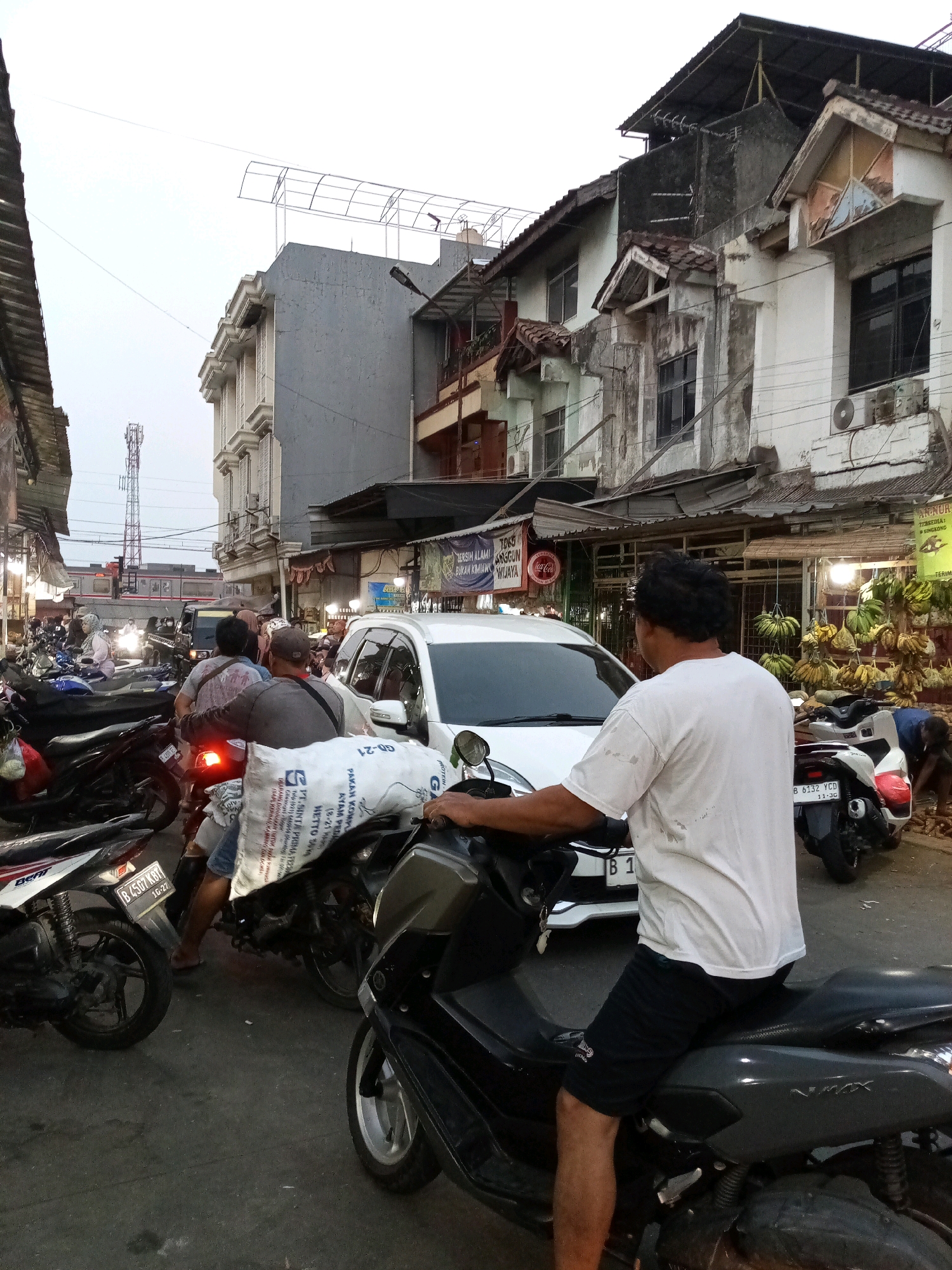 This image depicts a busy street scene in Bekasi, Indonesia. In the foreground, two men are riding motorcycles. The man in the center is wearing a white t-shirt and black shorts, while the man behind him is wearing a gray t-shirt and a cap. Both are on dark-colored scooters. The motorcycle in the foreground is a dark grey NMAX scooter, and the motorcycle behind it is a lighter colored scooter carrying a large white sack filled with what appears to be rice or grain.
Behind the motorcycles, a white car is visible, stopped in traffic. Further back, a marketplace or roadside stalls are set up, with bananas and other produce displayed. The buildings lining the street are multi-story, with some appearing to be shops or residences. A cell tower is visible in the distance. The sky is overcast, suggesting it is either early morning or late afternoon. The overall impression is one of a congested urban environment during daytime.