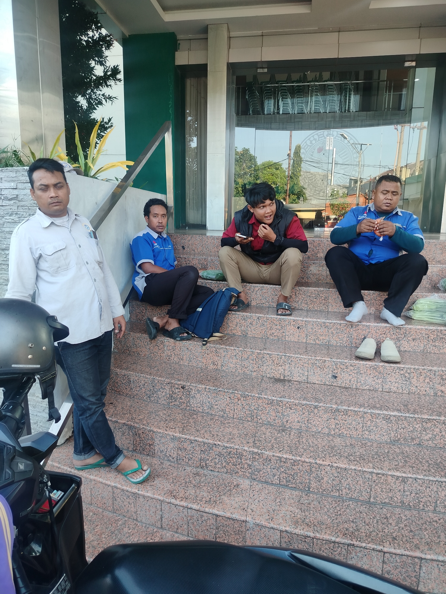 The image depicts four men seated on the granite steps outside a building, likely in Surabaya, Indonesia. The man on the far left stands and is partially obscured by a motorcycle. He wears a light-colored, collared shirt and dark jeans. The second man from the left sits on the steps, wearing a blue and white polo shirt and dark pants. A blue backpack is beside him. The third man, wearing a red vest over a darker shirt and tan pants, is seated and appears to be holding a phone. The fourth man from the left is also seated, dressed in a blue long-sleeved shirt and black pants, and is holding an orange packet. Green leafy vegetables are visible on the steps to his right. The background shows a glass-fronted building with stacked chairs visible inside. The overall setting suggests a casual outdoor waiting area or break spot.