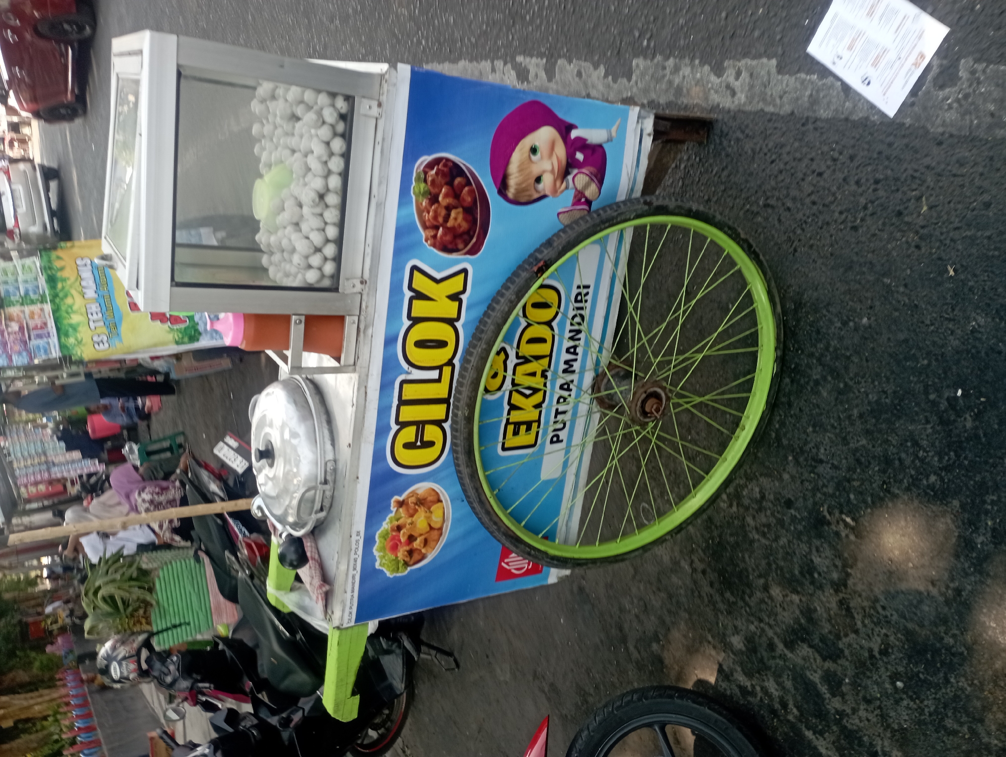 This is a medium shot, taken from a slightly elevated angle, of a food cart selling *cilok* (a type of Indonesian meatballs) on a street in Nganjuk, Indonesia. The cart is predominantly blue and white, with graphics of *cilok* and an illustration of the character Masha from the animated series *Masha and the Bear*. To the left of the cart are two parked motorcycles. In the background, there are several people, some sitting and some standing, along with other food vendors and parked cars. The ground appears to be wet asphalt, suggesting it may have recently rained. The overall impression is of a busy street scene with informal food commerce.