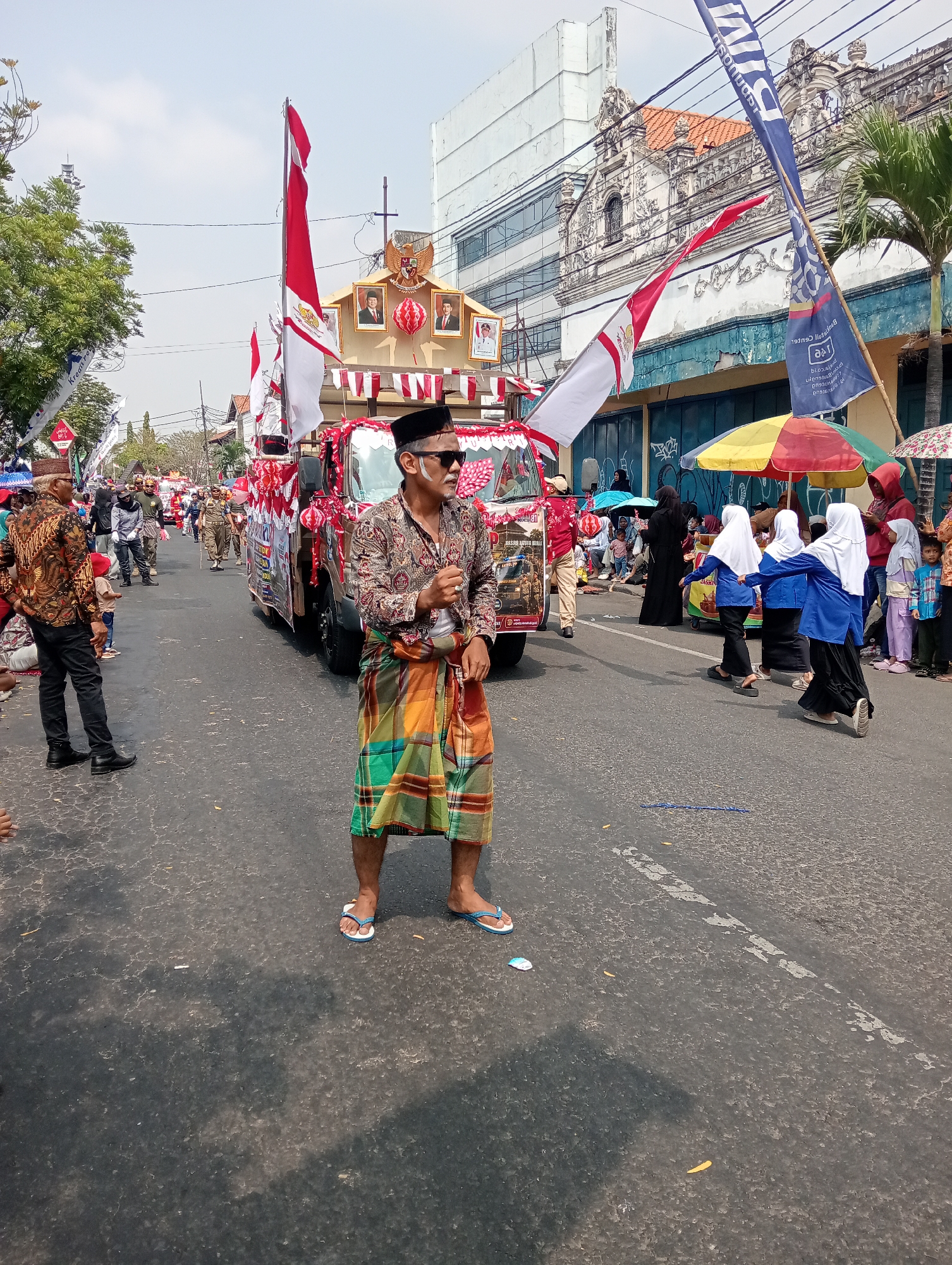 This is a photograph taken on a street in Demak, Indonesia, during a parade. In the foreground, a man wearing a patterned shirt, a sarong, sunglasses, and a black cap is standing in the middle of the road, holding something in his right hand. Behind him, a decorated truck is moving, adorned with flags, pictures of individuals, and red and white banners. Several people, including women in headscarves, are walking alongside or observing the parade. The street is paved, and buildings with distinct architectural features, including ornate facades and tiled roofs, line the background. The sky is clear and bright, suggesting it is daytime. Trees and palm trees are visible on the sides of the street. The scene depicts a festive or celebratory event.