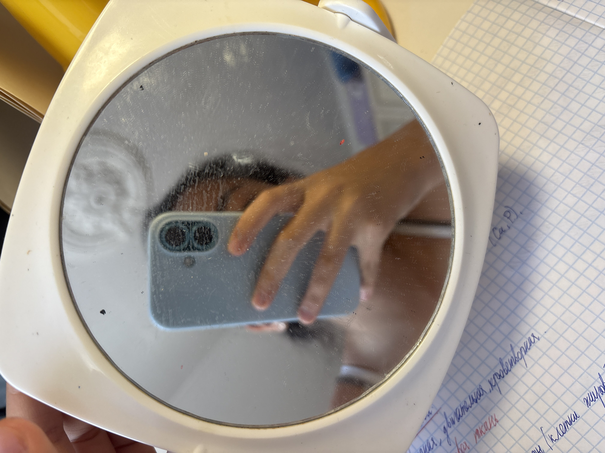 A person is holding a small, round mirror with a white plastic frame, taking a selfie with a blue smartphone. The mirror is reflective, showing the person's face, hand, and the smartphone. The background is blurred, but appears to be an indoor setting with papers and possibly a yellow object. There are some smudges and dust particles on the mirror's surface. A handwritten note on graph paper is visible in the foreground. The image does not contain any information that would indicate a specific location in Georgiyevsk, Russia.