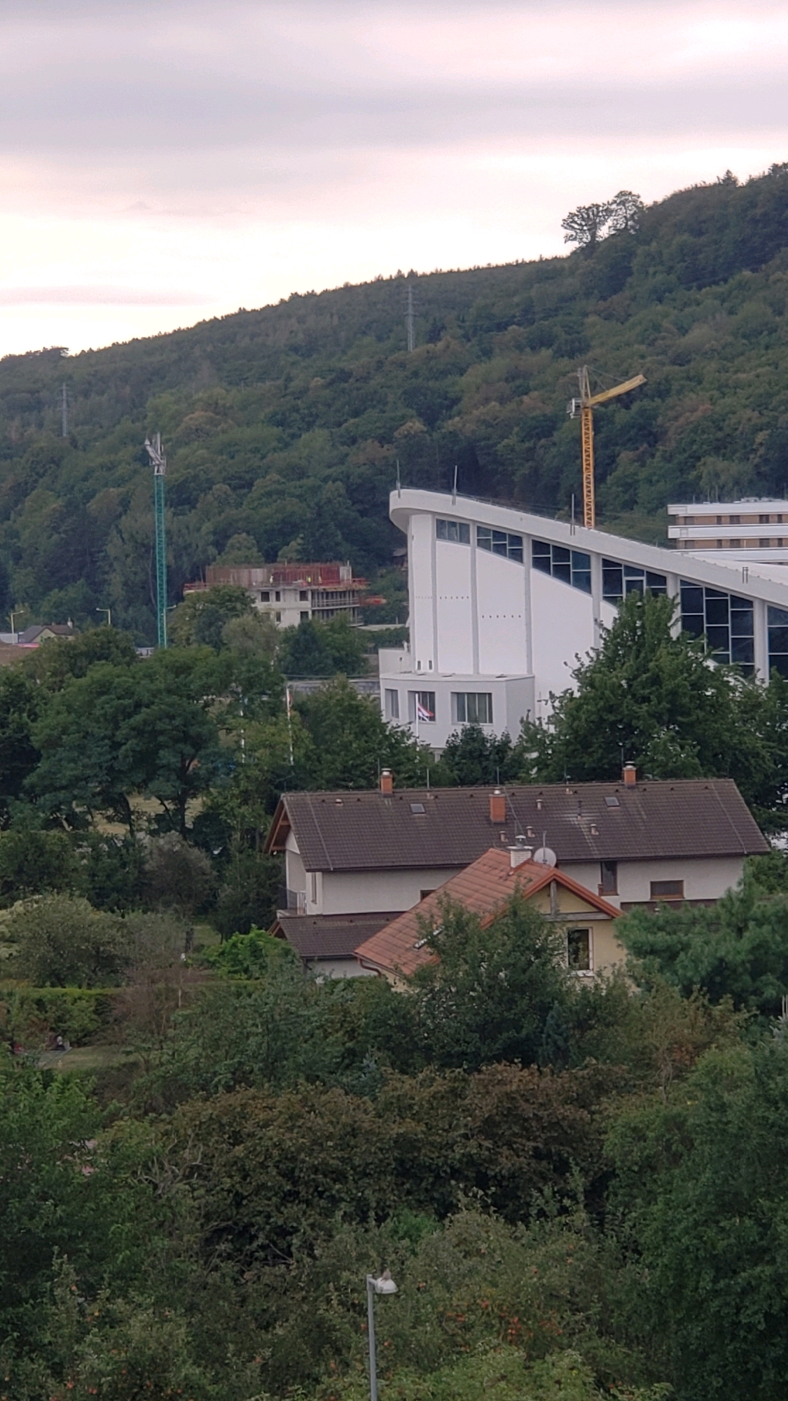 The image captures a view of the eastern side of the Slanské vrchy hills, with a portion of the city of Prešov, Slovakia, visible in the foreground and mid-ground. The overcast sky suggests it might be late afternoon or early evening.
Dominating the mid-ground is a large, modern white building with a distinctive slanted roofline, featuring numerous windows. To the left of this building, a construction site is partially visible with a tall green crane. Further in the background, on the hillside, are several communication towers and power lines.
In the foreground, several residential buildings with terracotta roofs are nestled among dense green trees and foliage. One building in the center has a prominent gable with a yellow facade and a visible window. Another building in the lower center is a two-story structure with a dark brown tiled roof and several chimneys. The overall scene is one of a semi-urban environment bordering a natural, forested landscape. No people are clearly discernible in the image.