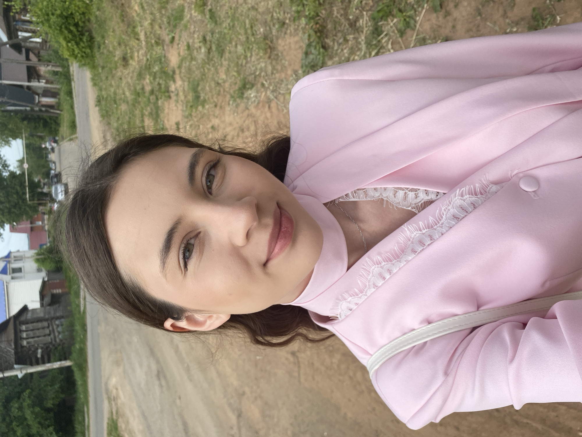 A young woman is outdoors, likely in Votkinsk, Russia, given the architectural style of the buildings in the background. She is wearing a pink blazer with white lace detailing and a white top underneath. The background consists of a grassy area, a road, and a glimpse of traditional wooden houses. There are cars visible in the distance, indicating a semi-urban setting. The woman appears to be looking directly at the camera with a slight smile.