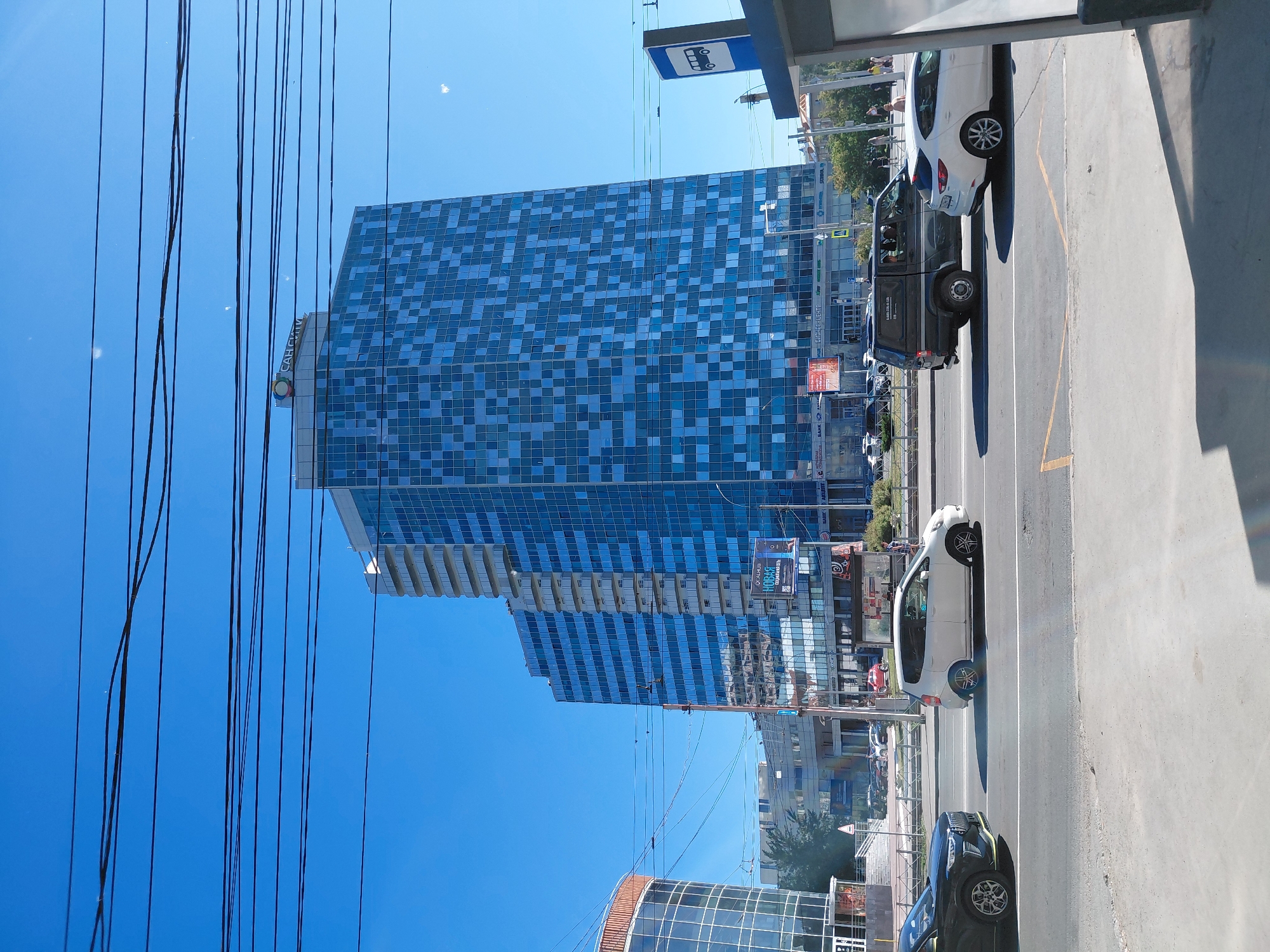 A wide shot captures a bright, sunny day with a clear blue sky, crisscrossed by a dense network of dark electrical wires. In the mid-ground, a large, modern skyscraper dominates the scene. Its facade is a striking pattern of blue and light blue glass squares, creating a checkered effect. The building has multiple setbacks, with balconies visible on the lighter sections. A circular logo with a stylized "S" inside it is perched on top of the building.
To the left of the skyscraper, a curved, glass-fronted building with orange accents is partially visible. Below the buildings, a multi-lane road is active with traffic. Several cars are on the road, including a black SUV and a white compact car in the foreground. A bus stop shelter with a blue sign depicting a bus is on the right side of the frame. In the background, other buildings and trees can be seen. The overall impression is a bustling urban environment under a clear, sunny sky.