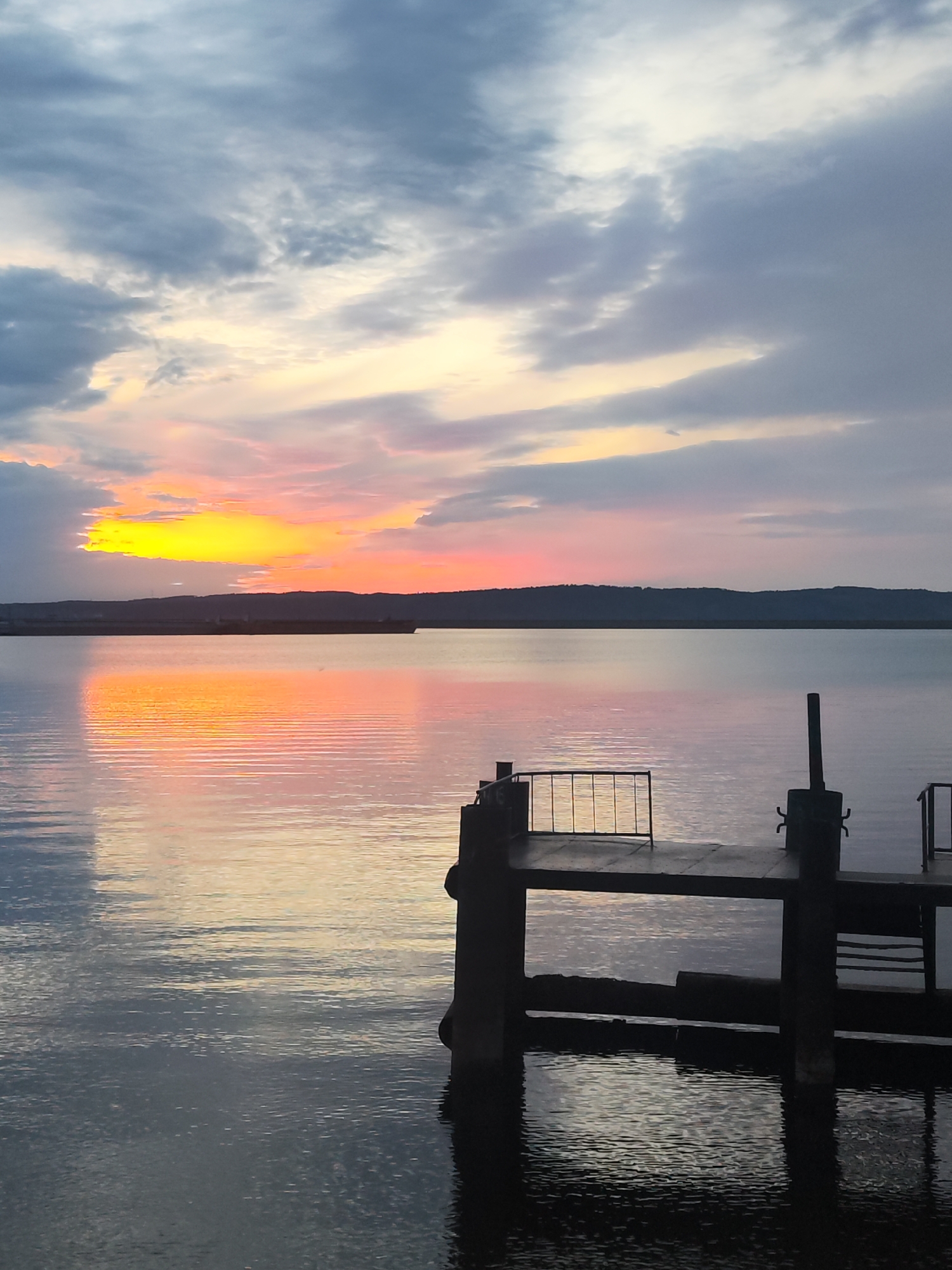 The image shows a sunset over the Kama Reservoir in Chaikovsky, Russia. The sky is filled with a mix of gray and orange clouds, reflecting in the water. A dark pier is in the foreground, partially submerged with reflections visible in the water. In the background, a line of trees and possibly a ship are silhouetted against the bright horizon.