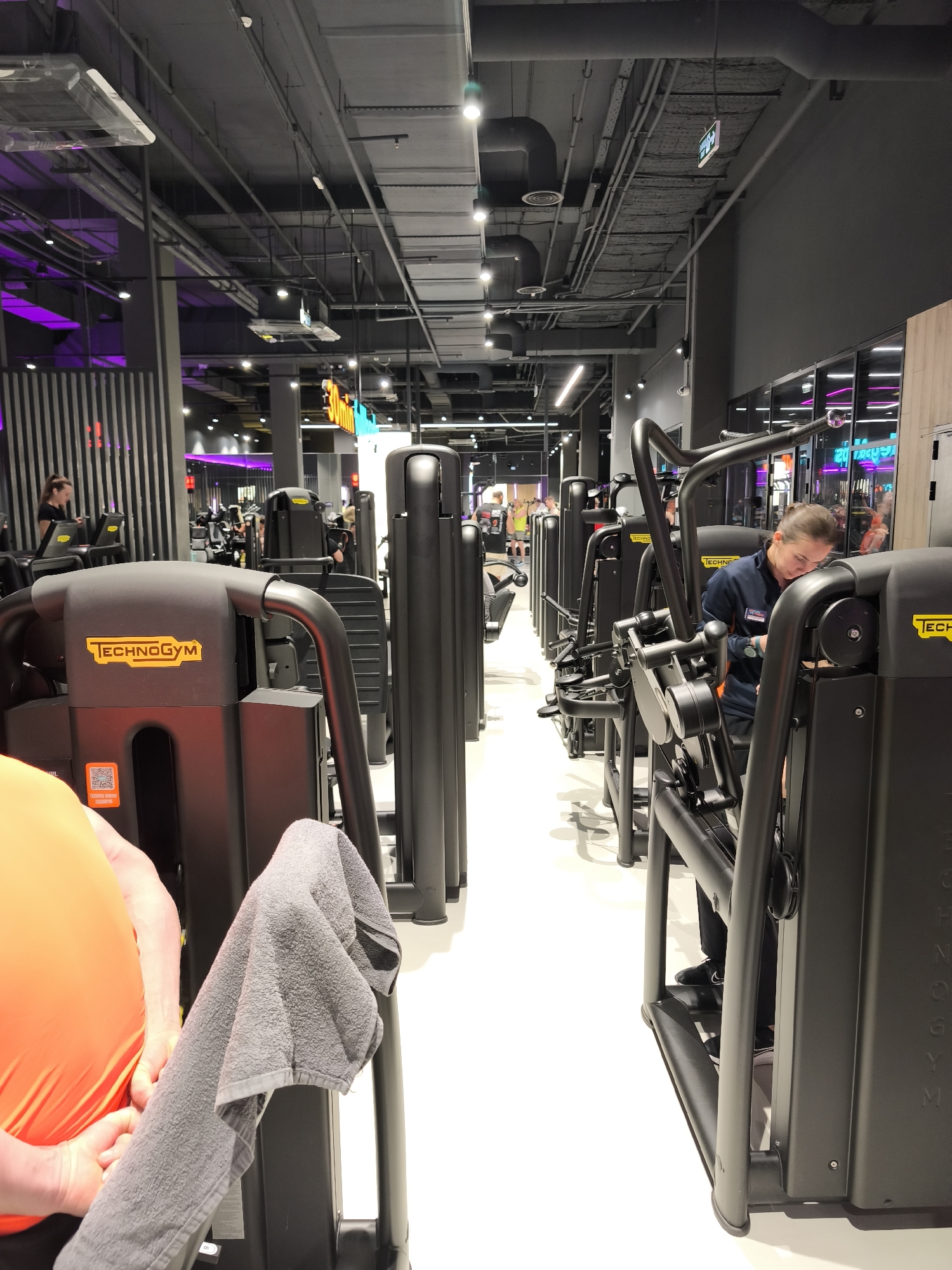 The image depicts the interior of a modern gym, likely located in a metropolitan area. The gym equipment, predominantly black and bearing the "TECHNOGYM" brand, is arranged in neat rows along a clean, white floor. Overhead, exposed pipes and ventilation systems run along the ceiling, which is painted in a dark gray color. Lighting fixtures are strategically placed throughout the ceiling, providing ample illumination.
Several individuals are present in the scene. A person wearing an orange top and holding a gray towel is partially visible in the foreground. Further down the line of equipment, a woman dressed in a dark blue jacket is interacting with one of the machines, possibly adjusting settings or cleaning it. In the background, other gym-goers can be seen engaging in various exercises.
The environment suggests a focus on fitness and well-being. The cleanliness and organization of the equipment contribute to the impression of a professionally managed space. The image appears to be taken in the city of Moscow, Russia.