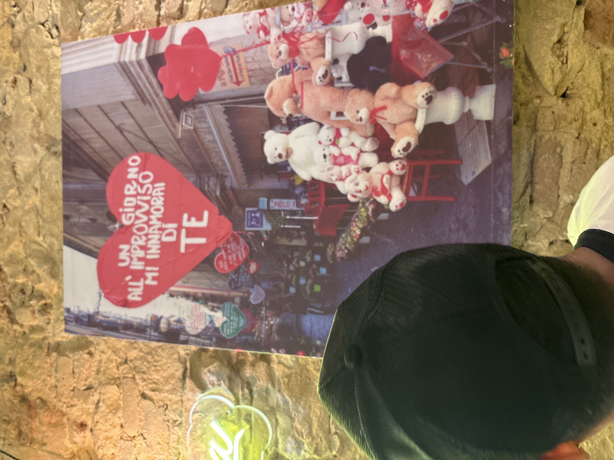 The image shows a close-up of a person wearing a black cap from behind, partially obscuring a poster. The poster depicts a street scene with numerous teddy bears displayed in front of a shop. Red heart-shaped balloons and decorations are prominent. Text on the poster reads "UN GIORNO ALL'IMPROVVISO MI INNAMORAI DI TE". The background of the poster shows a street lined with flower stalls. To the left of the poster, a portion of a neon light sign shaped like a cloud is visible. The entire scene appears to be indoors, with a stone wall forming the background behind the poster and the person. There is no information within the image to suggest the location is Barcelona, Spain.