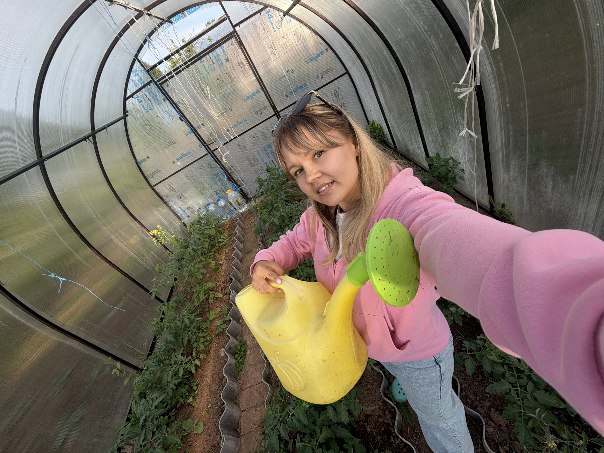 A young woman is captured in a selfie taken inside a greenhouse. She has blonde hair with bangs and wears sunglasses pushed up onto her head. Dressed in a light pink hooded sweatshirt and light blue jeans, she also has light blue Crocs-like shoes on her left foot. She is holding a vibrant yellow watering can with a bright green rose-spout in her right hand, poised as if about to water the plants. A gentle smile plays on her lips, suggesting a pleasant demeanor.
The scene is an arched, tunnel-shaped greenhouse constructed with transparent, corrugated plastic or polycarbonate panels and a dark metal frame. Rows of green plants, likely young tomato plants, are growing in the brown soil along the sides of a central pathway. Some blue and white strings are tied to the greenhouse structure above the plants, possibly for support. Visible through the translucent panels are glimpses of a bright, sunny sky and outdoor greenery, indicating it is daytime.
Several logos and text are visible on the greenhouse panels, including "Sellex", "14 years", "Inside", and "xajas", which appear to be branding or specifications for the greenhouse materials. The activity depicted is tending to plants, specifically watering them within this agricultural structure. The location context provided is Чумна, Russia.