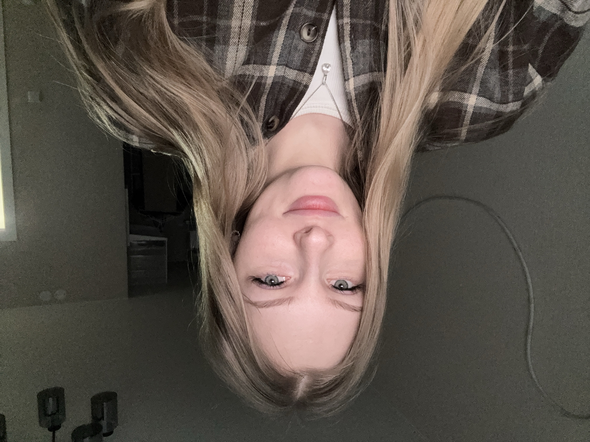 A young woman with blonde hair is seen upside-down in the frame, wearing a brown and white plaid shirt over a white top with a silver necklace. Her eyes are light blue, and her complexion is fair. The background is dimly lit, revealing a glimpse of a room with what appears to be furniture and a window. A coiled cable is partially visible on the right side of the frame. The location of the image within the city of Perm, Russia, is not discernible from the given details.