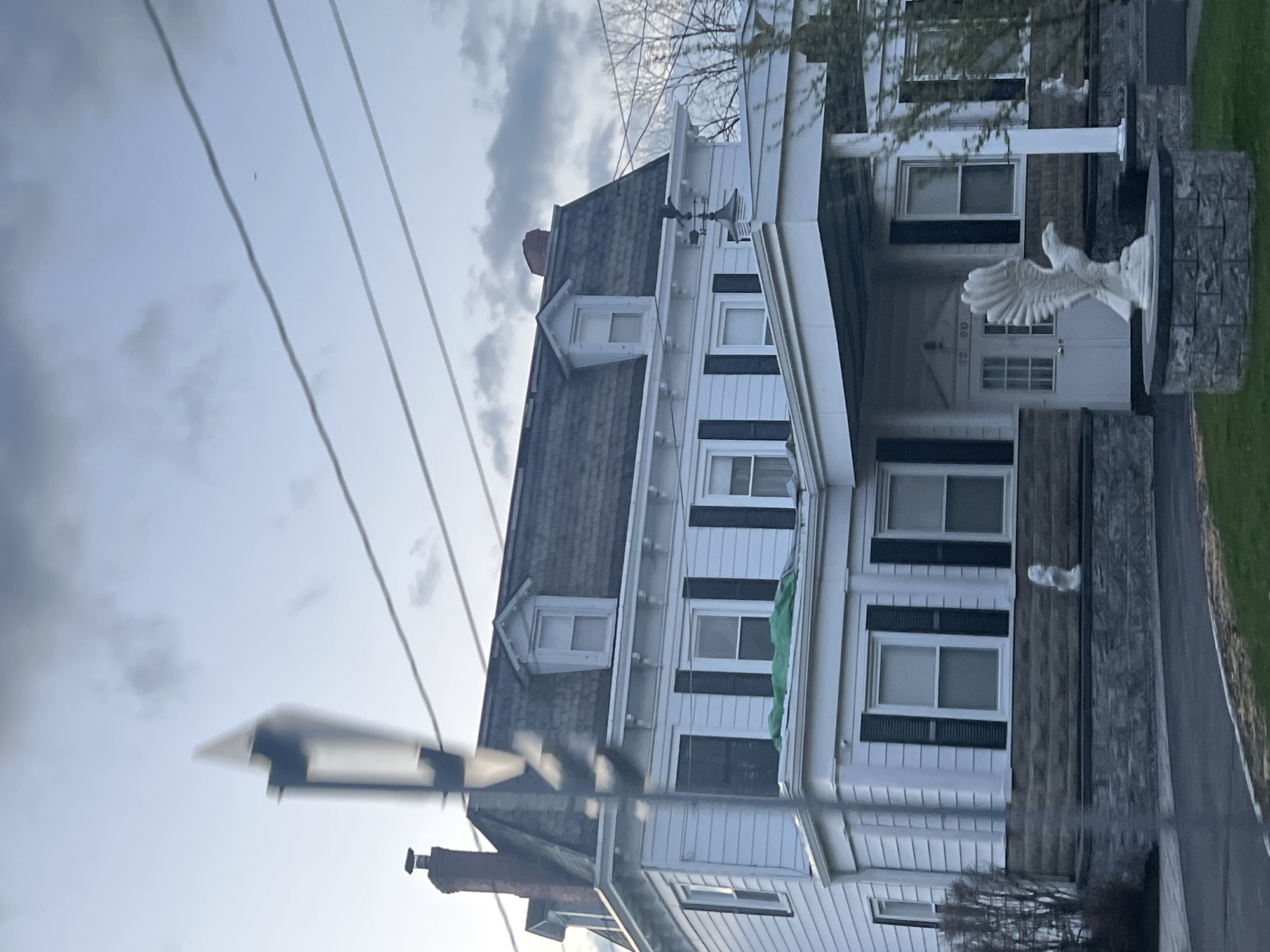 The image depicts a white, multi-story house with black shutters and a gray shingled roof. A prominent white sculpture of an eagle with outstretched wings stands in front of the house on a stone pedestal. Overhead power lines are visible in the foreground against a cloudy sky. The house number "1290" is visible above the door. The photo was taken in Marlboro, United States of America.