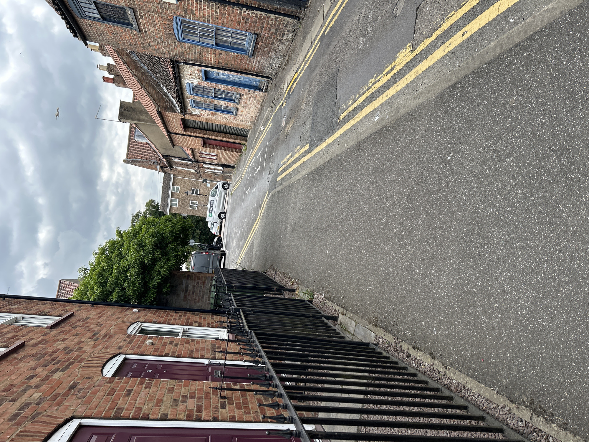 This is a slightly tilted, eye-level outdoor shot of a narrow street in Boston, United Kingdom. The scene depicts a residential area with brick buildings lining one side of the asphalt road. The road has double yellow lines painted along its center.
On the left side of the street, we see the facades of brick houses. The lower buildings have burgundy doors and white-framed windows. Further up the street, older brick buildings with blue-framed windows and exposed brick are visible, suggesting a mix of architectural styles and ages. A large, leafy green tree stands between the older and newer buildings, its branches reaching towards the sky.
On the right side of the road, a black metal fence runs along the property line of the brick houses, separating them from the pavement. Beyond the fence and along the street, a few vehicles are parked, including a silver van with "enterprise" branding visible.
The sky is overcast with heavy grey clouds, and a lone bird is in flight against the cloudy backdrop. The overall impression is a slightly damp, perhaps cool, day in a historic urban environment. There is no discernible activity or event taking place, suggesting a still moment in time. The image is characterized by its brick textures, the contrast between the weathered old buildings and the newer ones, and the starkness of the overcast sky.