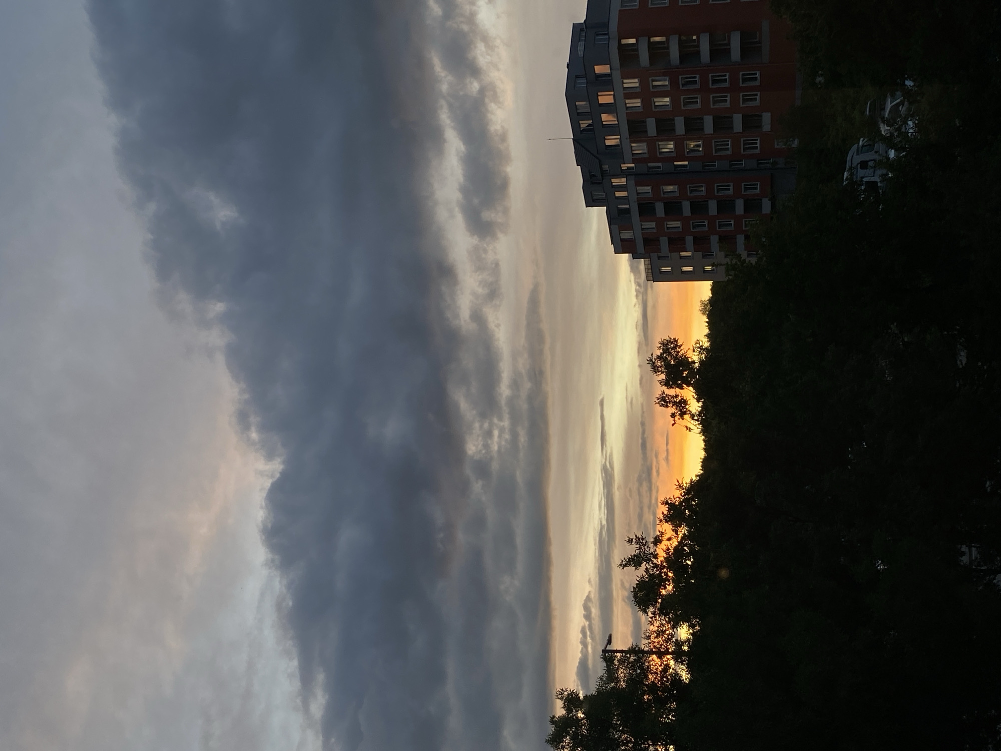 A wide-angle, low-angle, outdoor shot captures a dramatic sunset over a city skyline. Dark, heavy clouds dominate the upper portion of the frame, with lighter streaks of orange and yellow peeking through from the horizon. The warm glow of the sunset reflects off the windows of a multi-story, modern apartment building situated on the right side of the image. In the foreground, the dark silhouettes of trees and foliage obscure the lower part of the scene, creating a sense of depth. A streetlamp with a single light fixture is visible on the left, silhouetted against the sky. A white van and other vehicles are partially visible amidst the trees in the lower right portion of the image. The overall impression is one of a city preparing for nightfall, with the natural beauty of the sunset contrasting with the man-made structures.