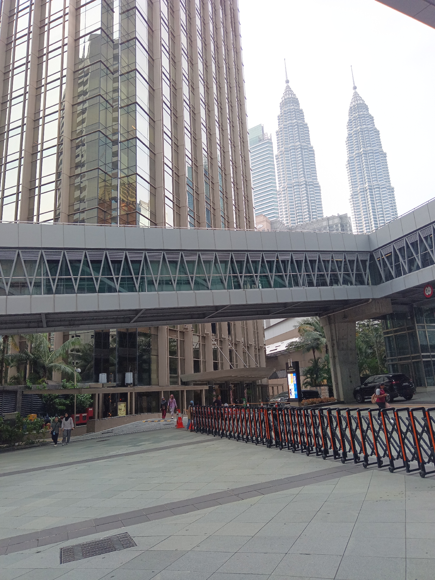 The image captures a wide-angle view of the area surrounding the Petronas Twin Towers in Kuala Lumpur, Malaysia. In the foreground, a grey tiled plaza is visible, with a metallic drain grate in the lower left quadrant. A long line of black and orange portable barricades stretches across the plaza, suggesting a controlled or cordoned-off area.
Behind the barricades, a modern elevated walkway with a geometric glass facade connects two buildings. This walkway is supported by grey concrete pillars. On the left side of the image, a tall, glass-fronted building with vertical metal accents dominates the scene, its facade reflecting the surroundings. On the right side of the walkway, more trees and the entrance to a building are visible.
In the middle ground, several individuals are walking on the plaza. On the far left, two women walk away from the camera. Further to the right, near the entrance of a building, a woman in a pink headscarf and a child are walking, while a woman in a grey headscarf and a man walk towards the camera. A black car is parked near the entrance on the right.
In the background, the iconic Petronas Twin Towers, with their distinctive spires, rise majestically against a bright, slightly hazy sky. Another modern skyscraper is partially visible to the left of the Twin Towers. The environment appears to be a bustling urban setting, likely a public space designed for pedestrians and visitors.