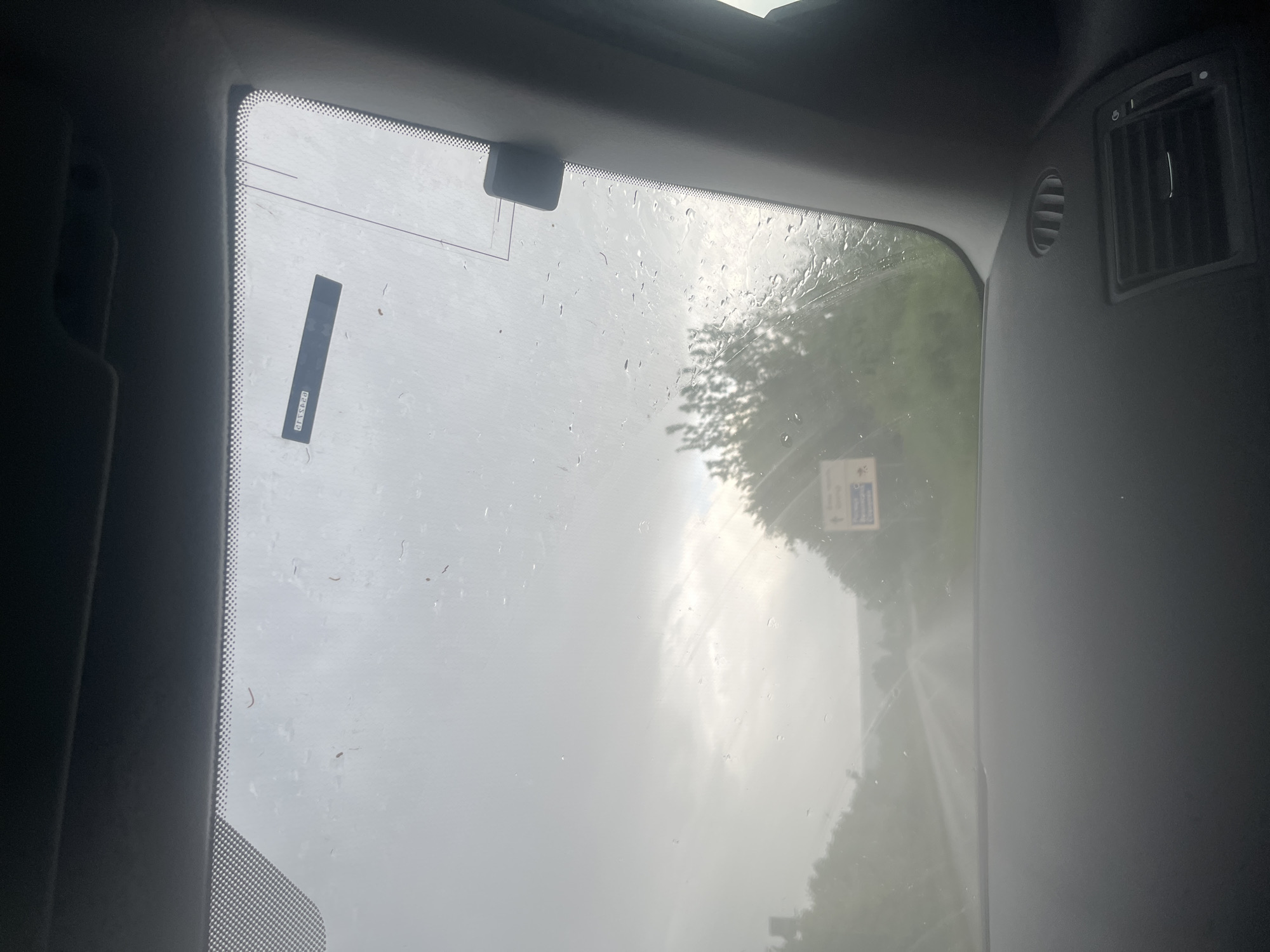 The image shows a view through a car window on a rainy day. A rectangular sticker is adhered to the windshield, with text visible, but illegible. A small, rectangular object is attached to the top of the windshield. Outside the window, trees and a road are visible, obscured by the rain and mist. A sign is vaguely visible on the side of the road, its text unreadable. The view suggests a road in a rural or suburban area during overcast weather. The image offers insufficient data to specifically identify a location in the city of Byala Cherkva, Bulgaria. An air vent is visible in the interior of the car to the right of the window.
