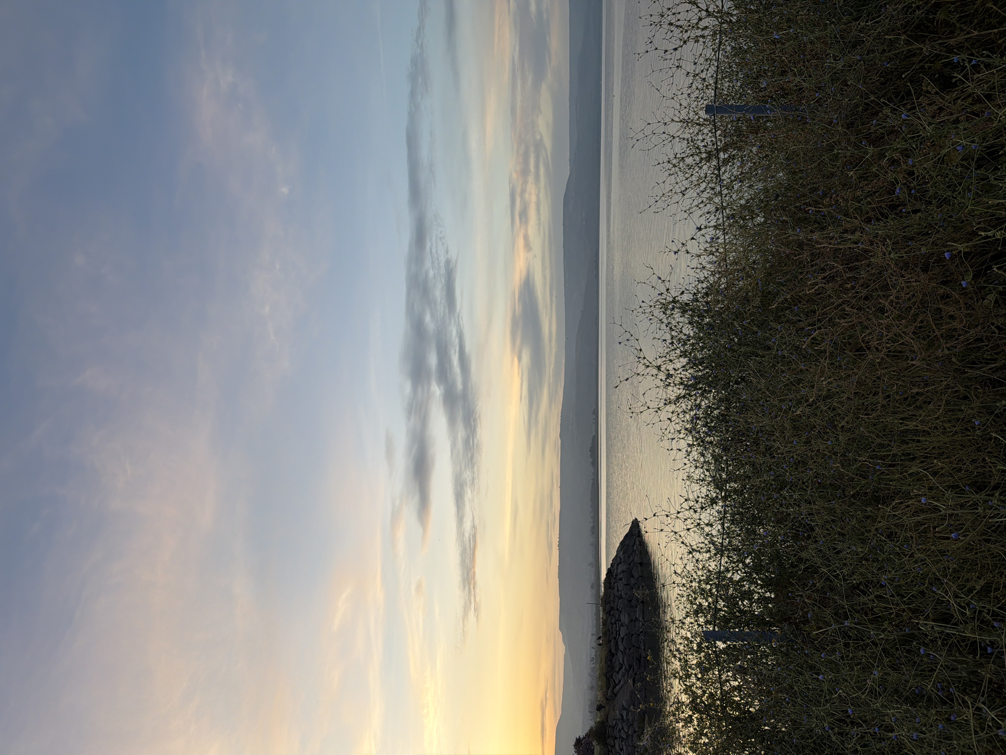 The image shows a landscape at the city of Emek HaYarden Regional Council, Israel. The sky is a gradient of light blue and yellow, transitioning into orange at the bottom. There are thin, vertical clouds in the center of the sky. A long body of water, possibly a lake or sea, stretches horizontally across the middle, with a distant shoreline and mountains visible on the opposite side. A pile of rocks and vegetation are visible on the right side of the image. The image is vertically oriented.